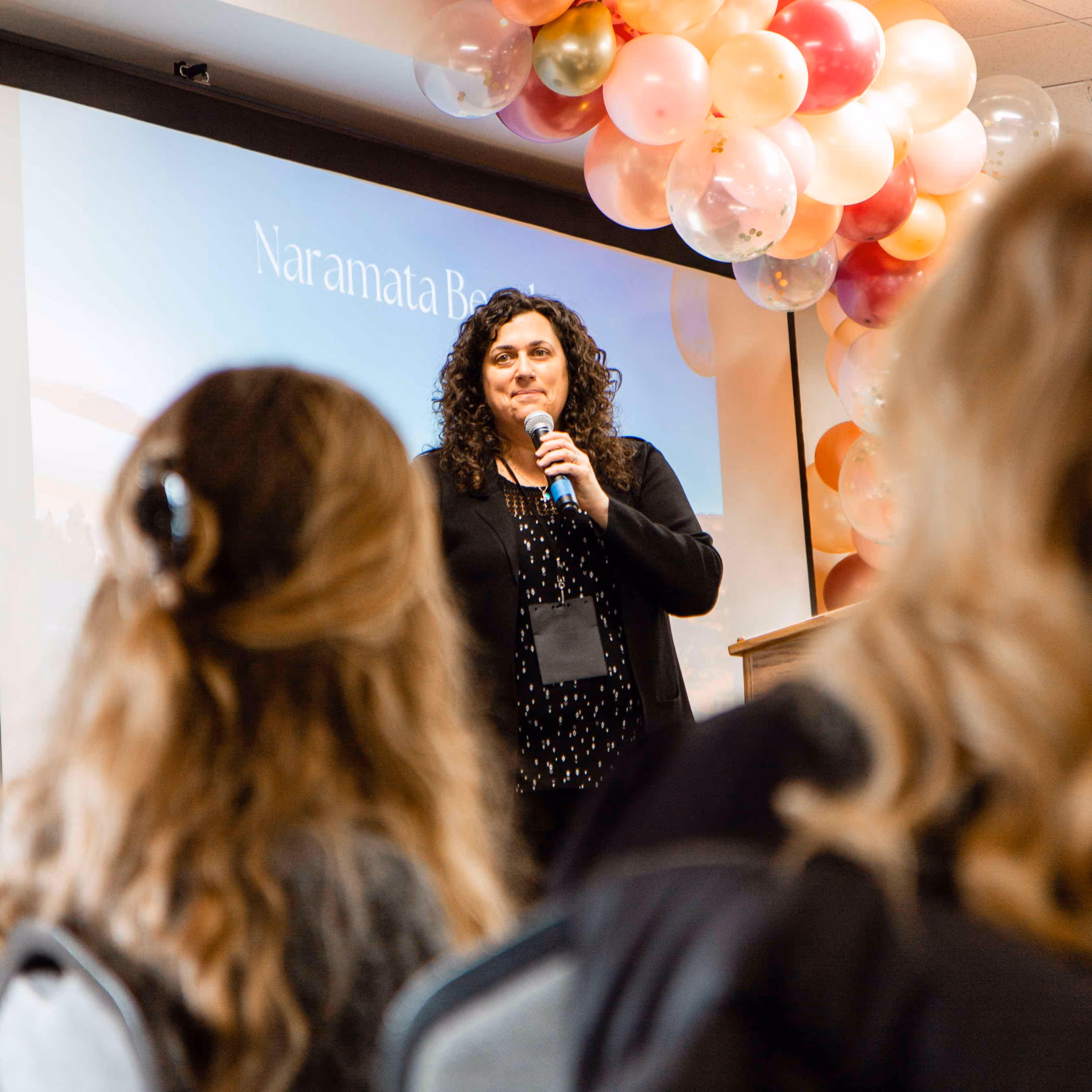 Woman speaking into a microphone at an event with a women in the foreground.