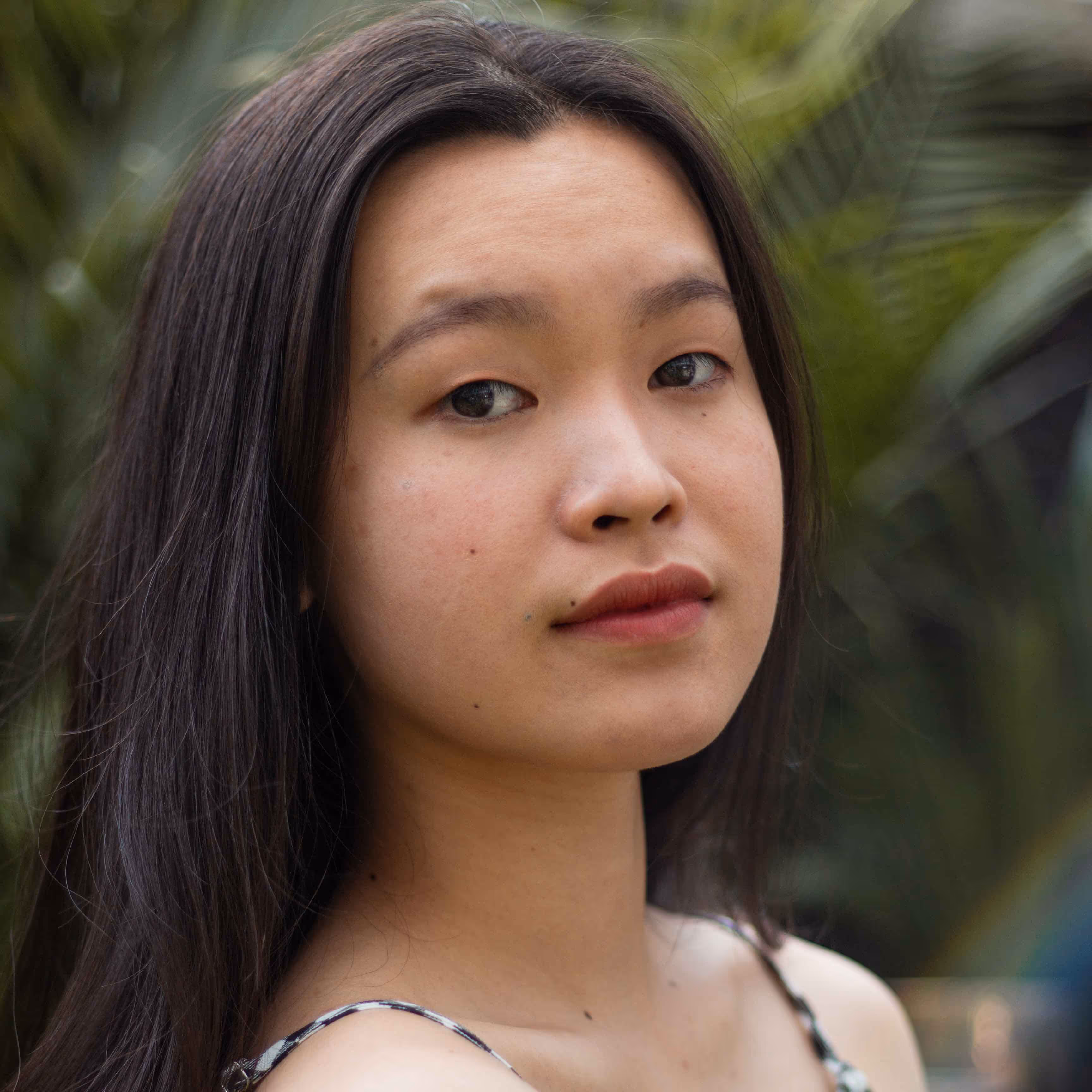 Close-up of a young woman with long dark hair and a neutral expression, standing in front of green foliage.