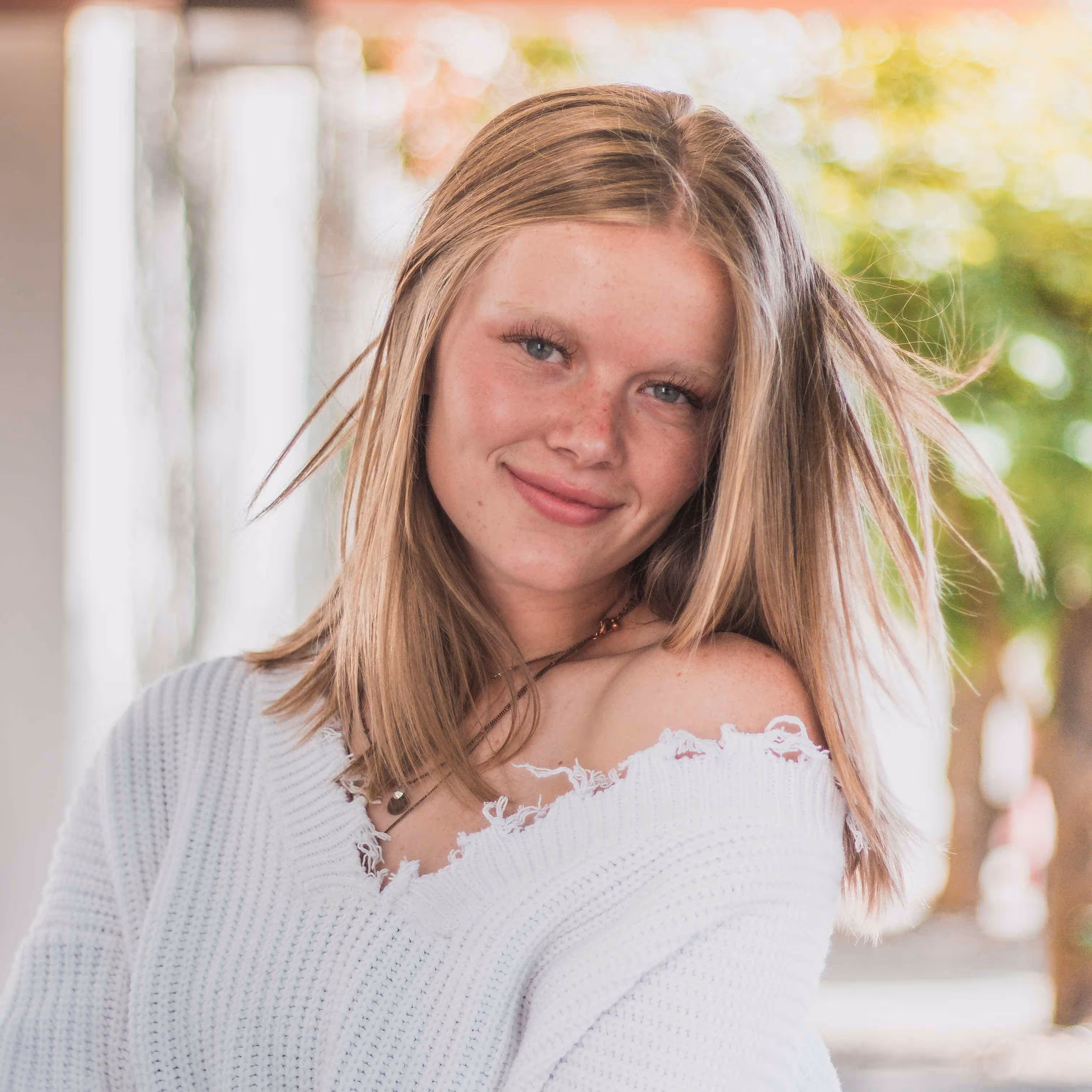 Smiling young woman with blonde hair wearing a white off-shoulder sweater outdoors.