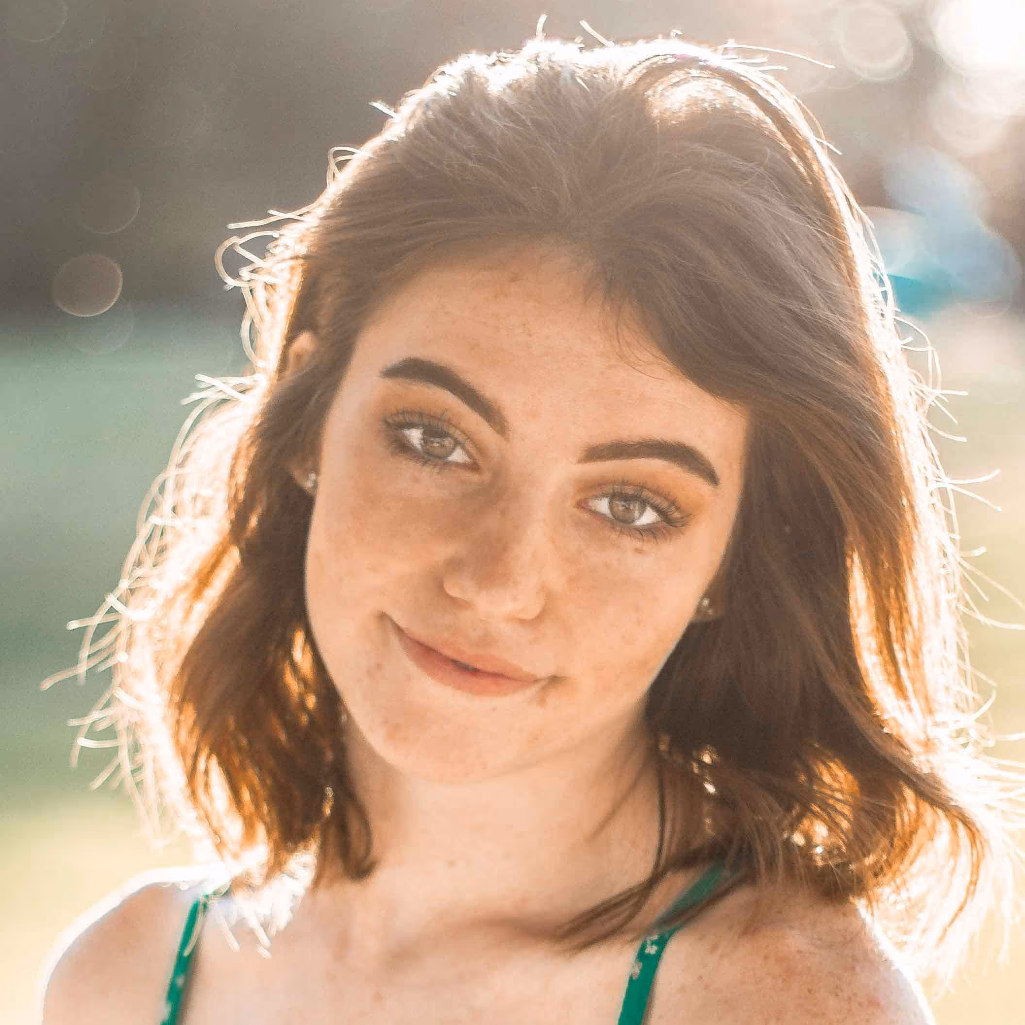 Young woman with shoulder-length brown hair smiling softly outdoors with sunlight behind her.