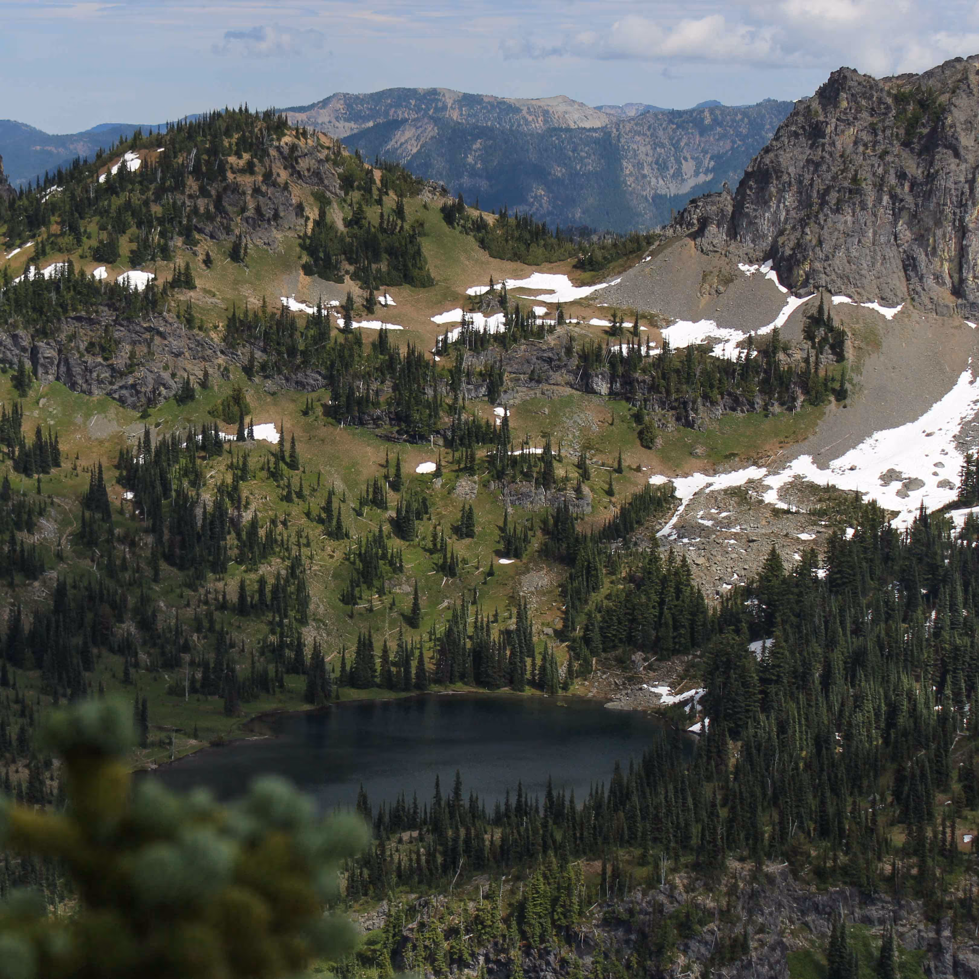 Mountain landscape with patches of snow, evergreen trees, and a dark alpine lake at the center.