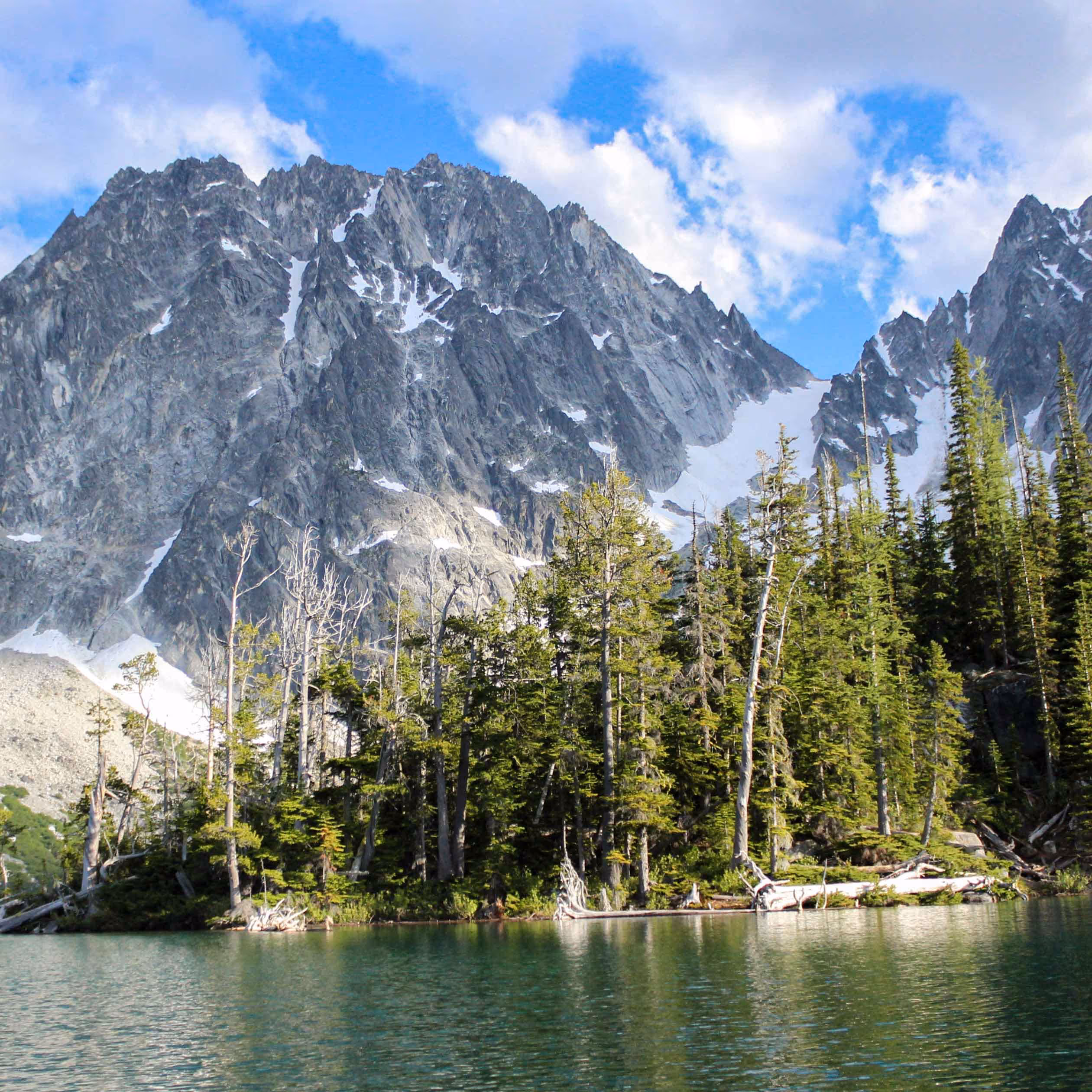 Clear alpine lake and rugged snow patches on tall mountain peaks under a partly cloudy blue sky.