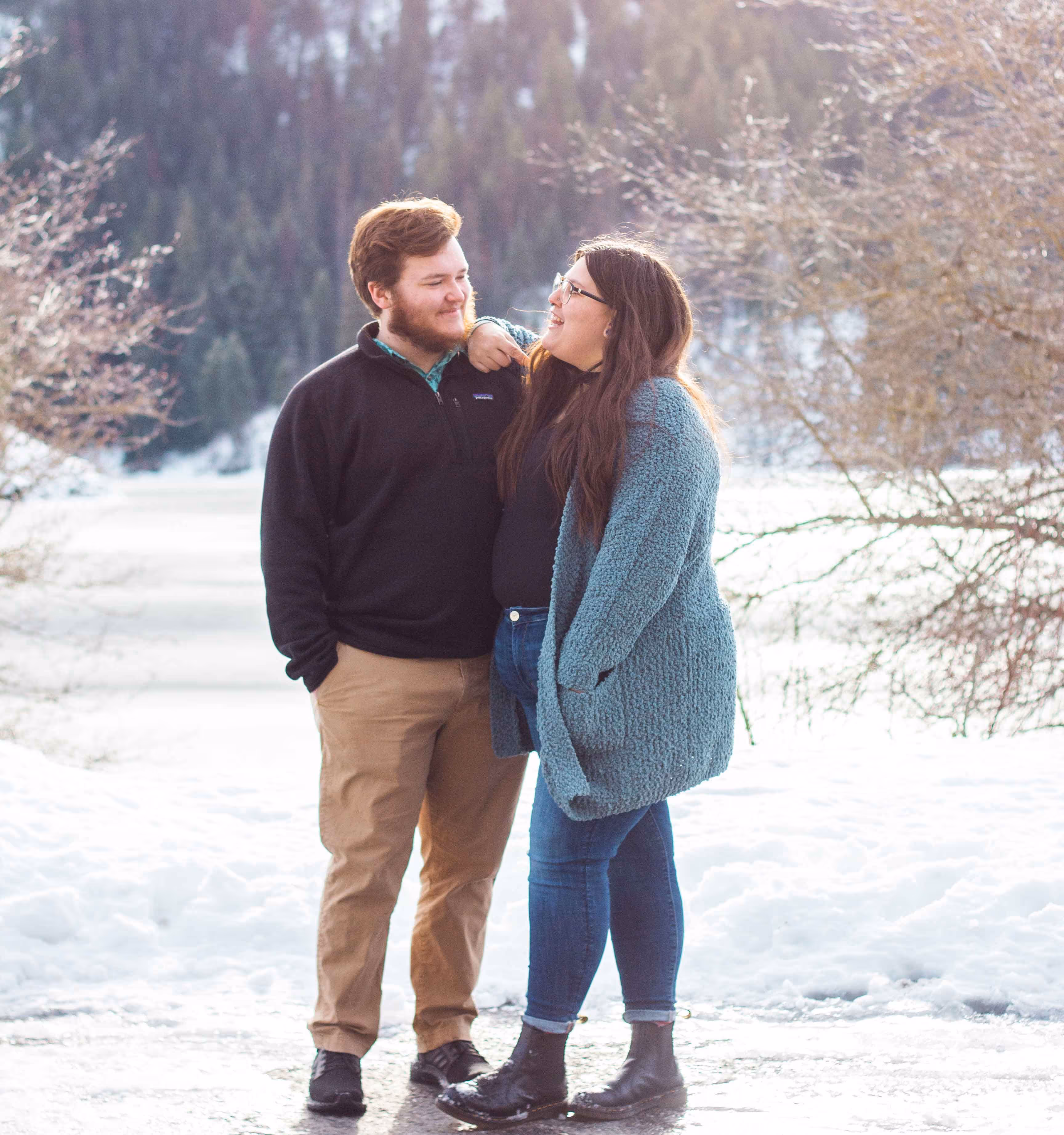 Couple dressed warmly standing on a snowy path, smiling and looking at each other with trees in the background.