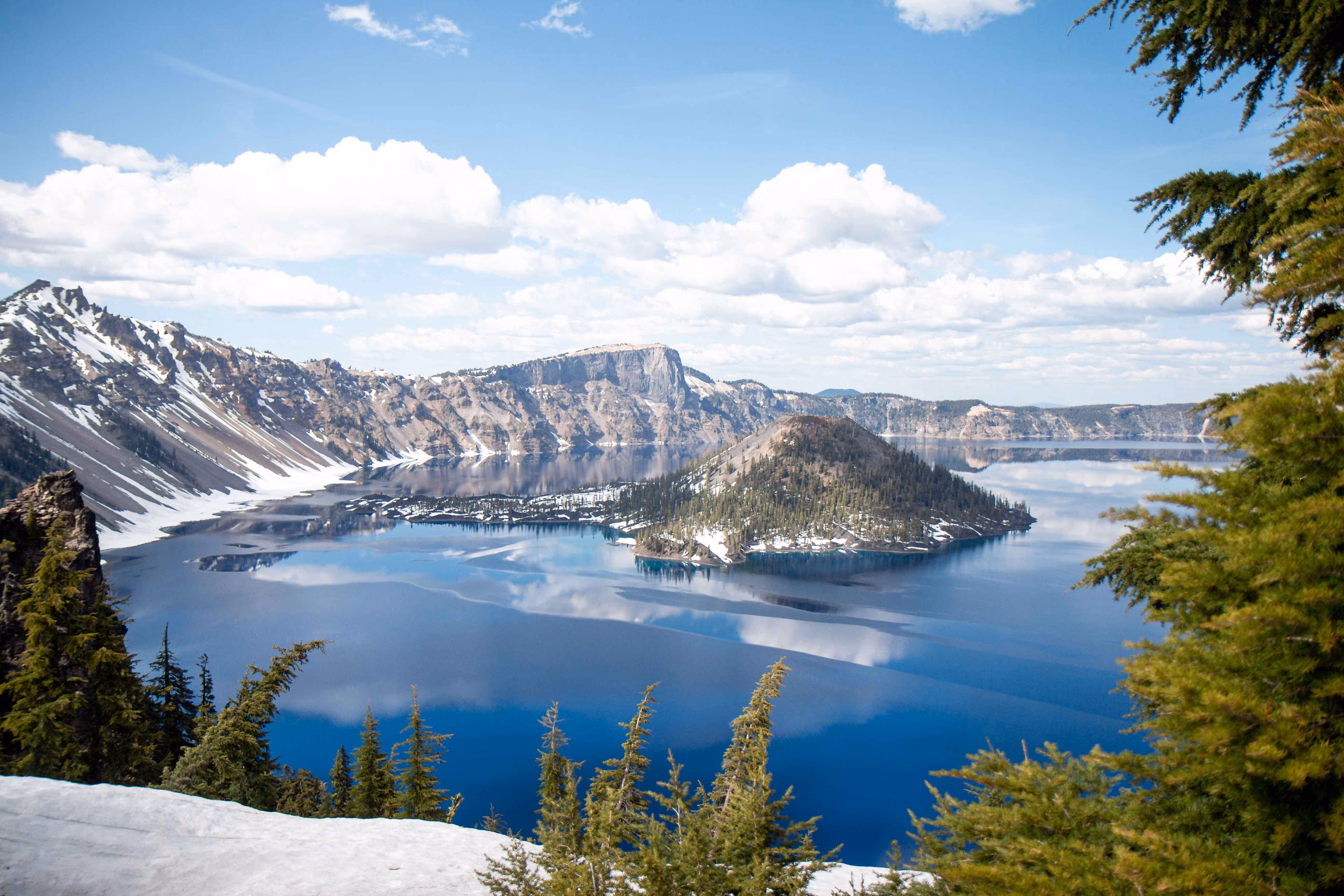 Clear blue crater lake surrounded by snow-capped mountains and evergreen trees under a partly cloudy sky.