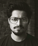 Black and white portrait of a man with glasses and a beard against a brick wall background.