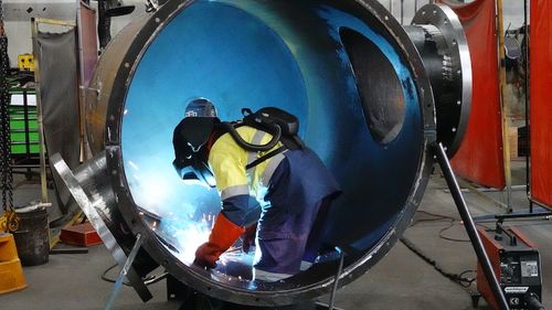 An industrial worker cutting a large metal tube and creating sparks.
