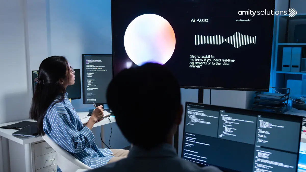 Two professionals in a dimly lit office reviewing an AI Assist interface on a large screen displaying a voice waveform and chat response, with code visible on surrounding monitors. Amity Solutions logo in the top right corner.