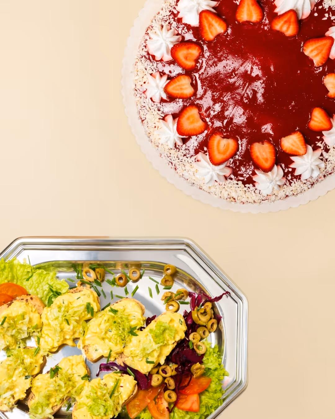 Erdbeertorte mit veganer Sahne und Gelee, darunter eine silberne Platte mit veganem Eiersalat auf Brot, garniert mit Oliven Salat und Tomaten.