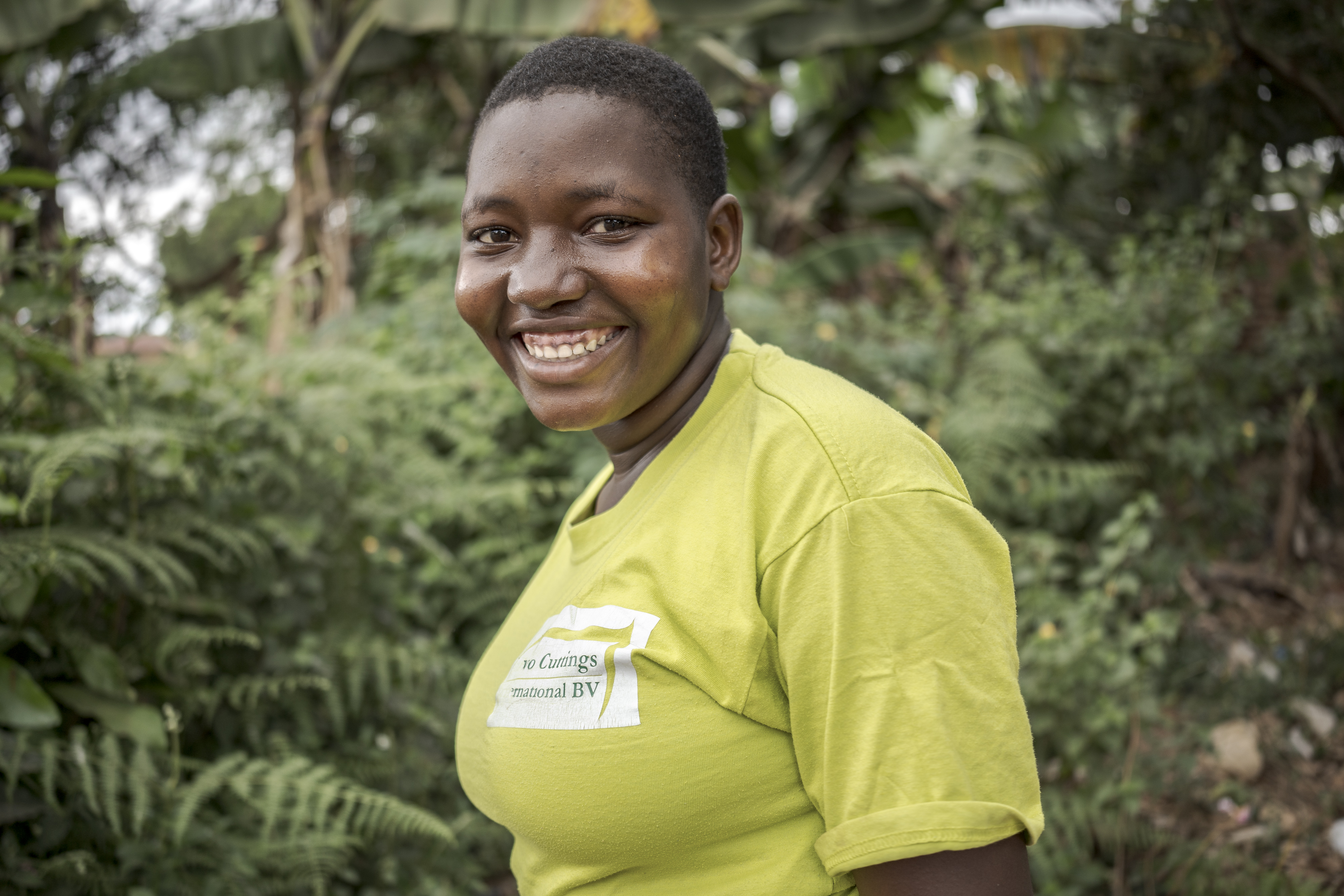 A teenaged girl in a yellow t-shirt smiles at the camera in front of a background of trees.