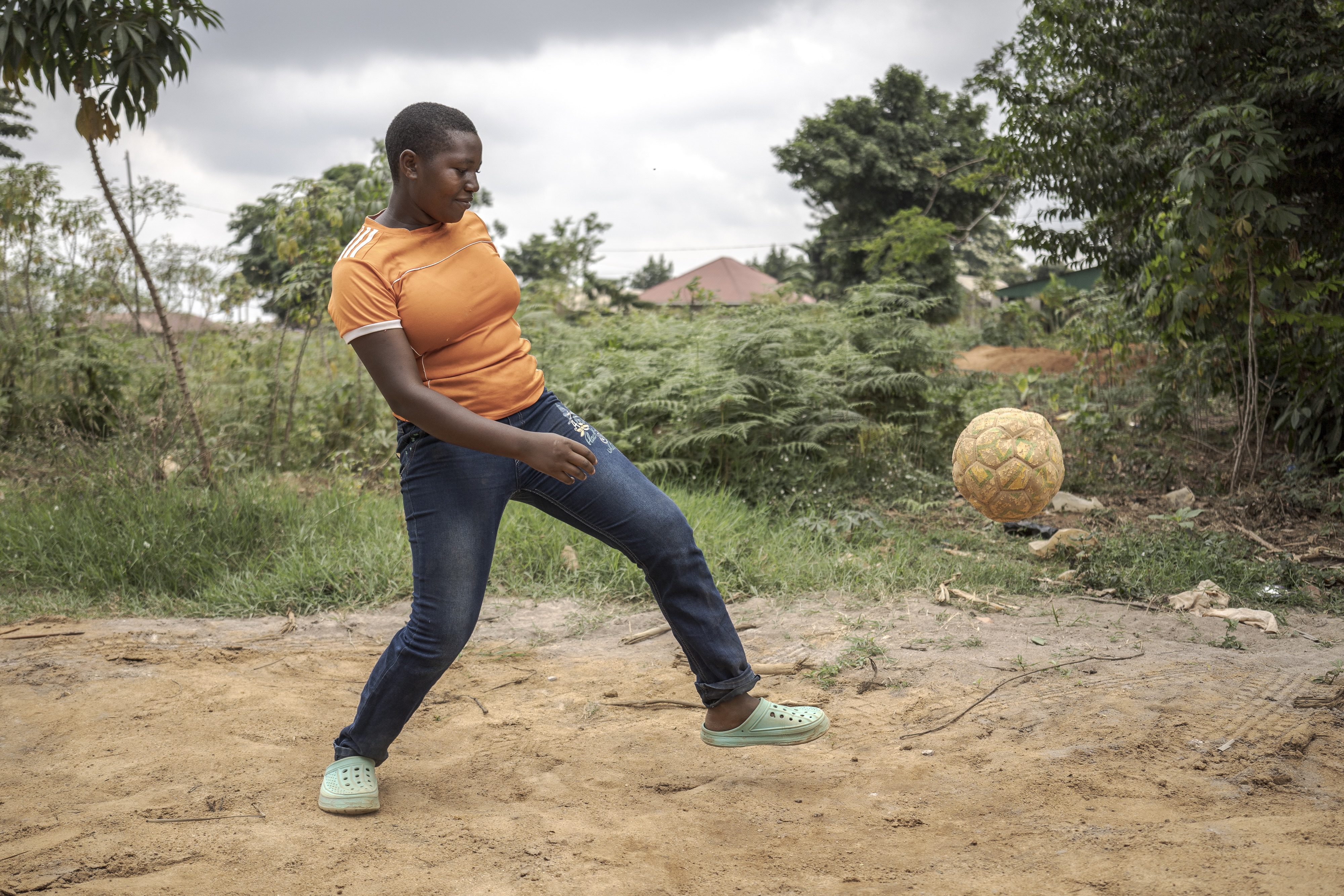 A teenaged girl in an orange t-shirt and blue jeans kicks a soccer ball.