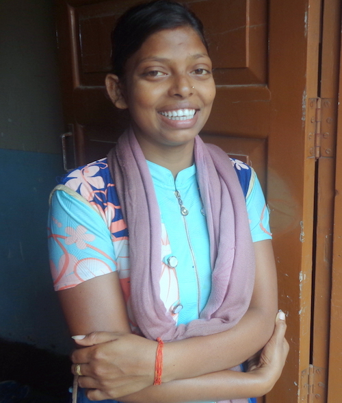 A girl with dark hair wearing a blue top with pink flowers and a pink scarf smiles into the camera while leaning on an open door.