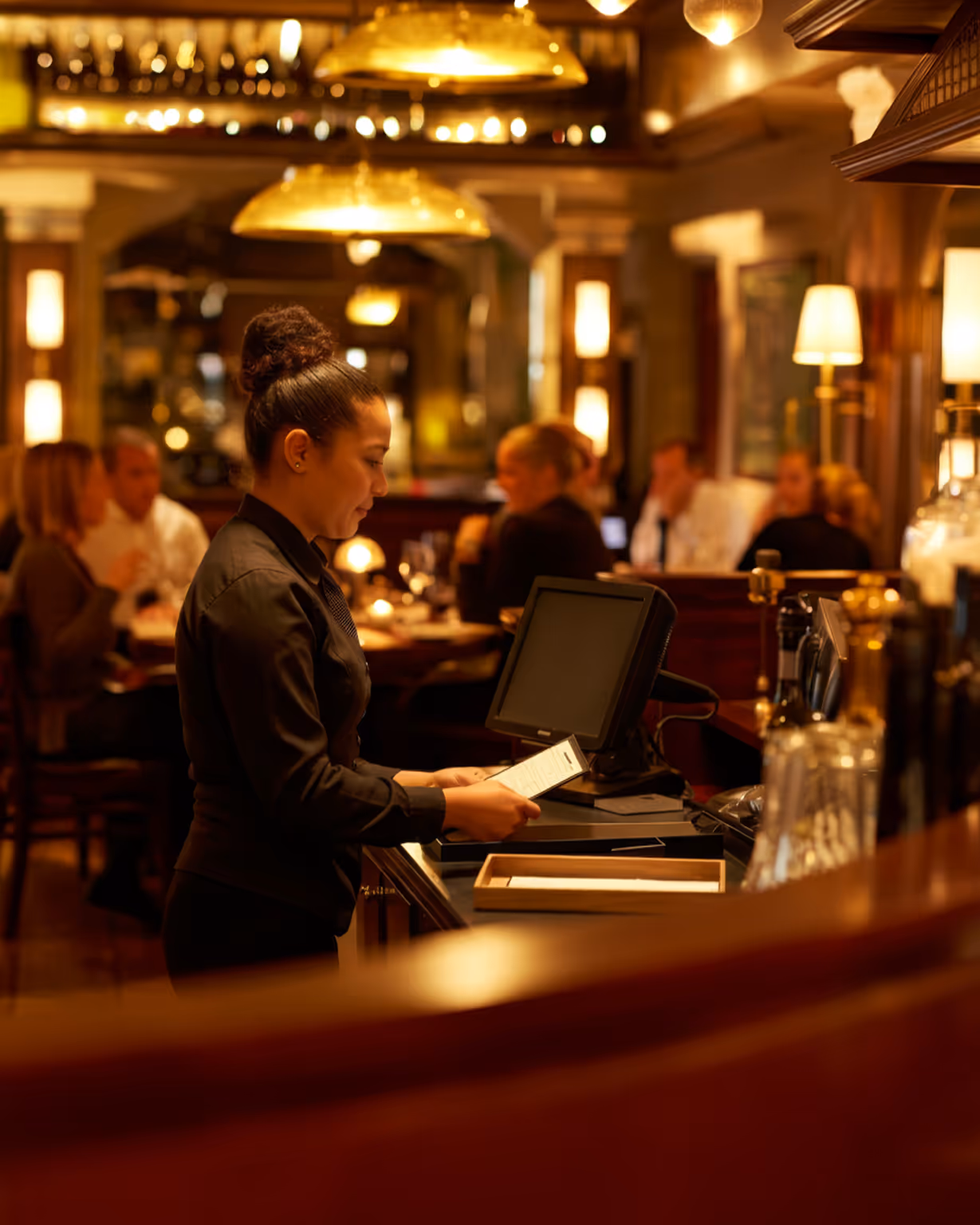 Waitress in black uniform reviewing a receipt at a restaurant counter with diners in the background.