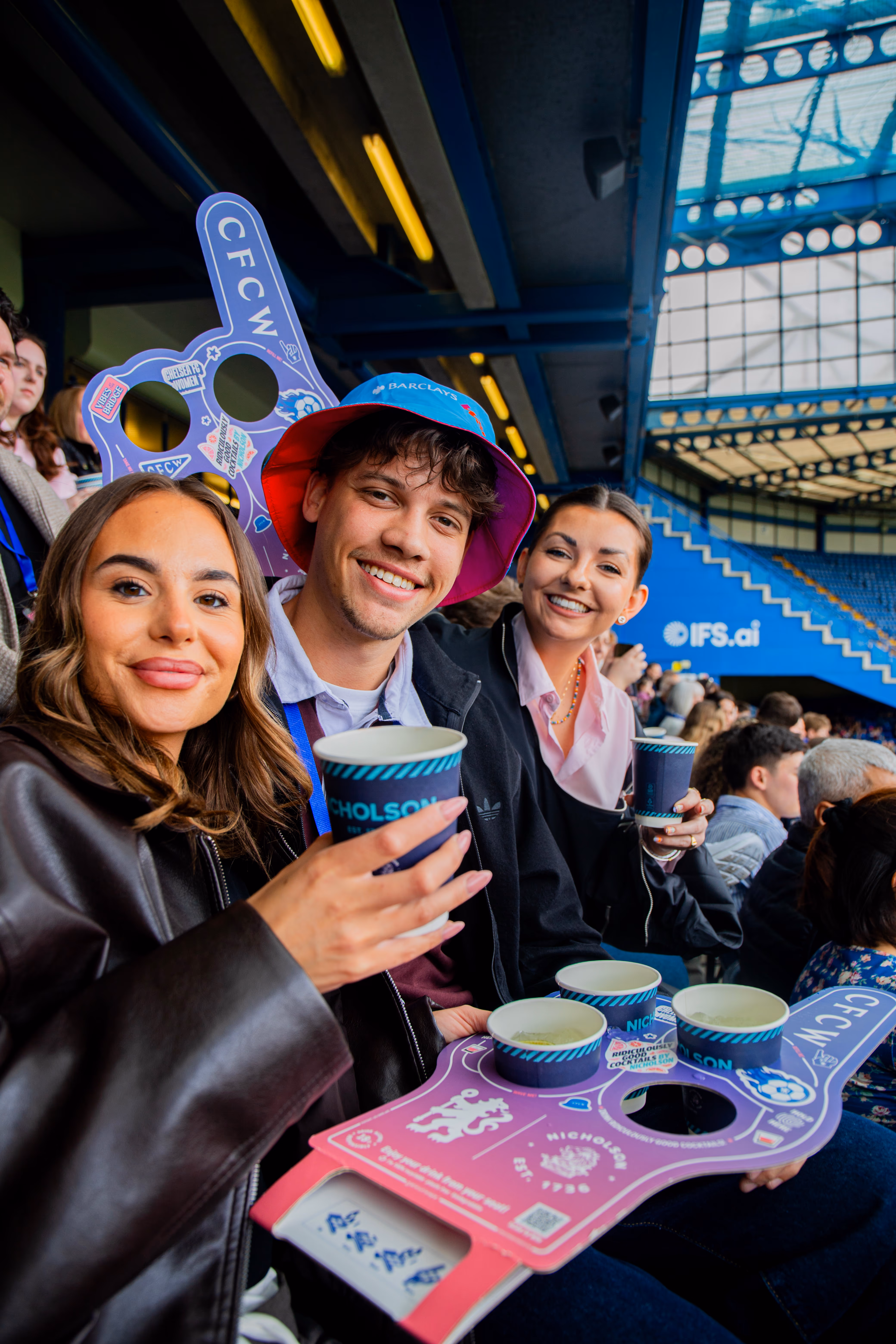 Three smiling fans hold drinks and a foam finger while sitting in the stands at a stadium, with rows of spectators visible behind them.