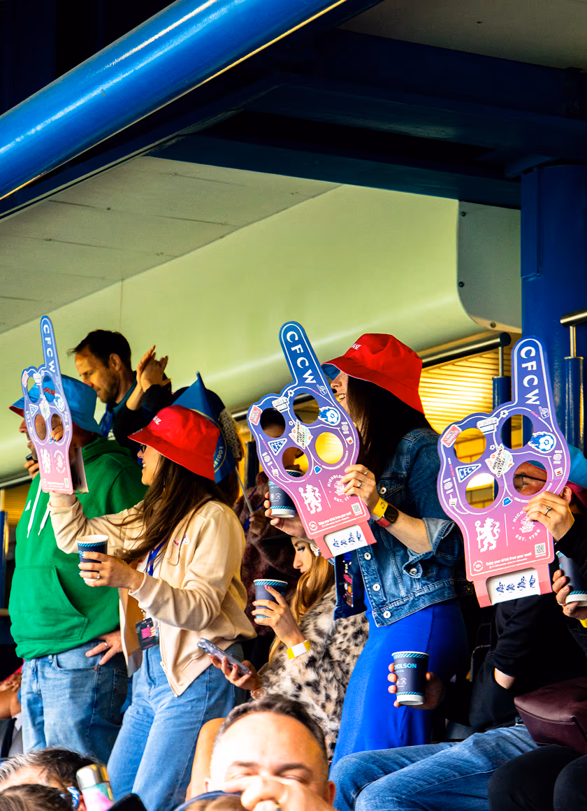 Fans at a Chelsea FC Women's stadium event hold up pink and purple foam finger cheer props with 'CFCW' printed on them.