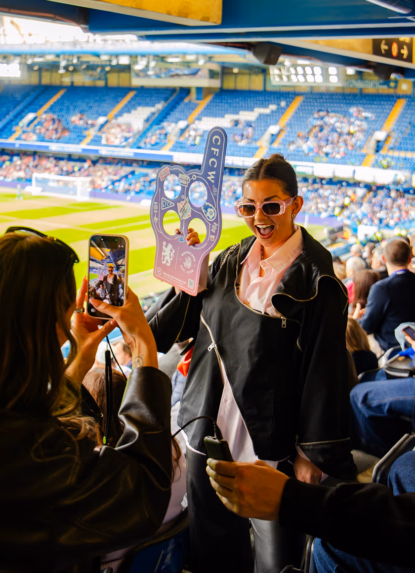 A woman wearing pink sunglasses and a black jacket smiles while holding a foam hand sign at a stadium, as another person photographs her with a smartphone.