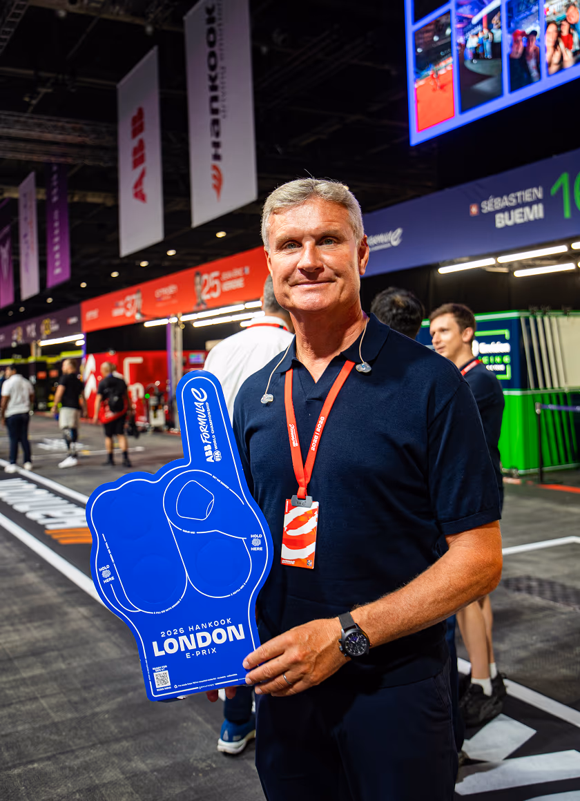 A smiling man in a navy polo shirt holds a large blue foam number-one hand promoting the 2026 Hankook London E-Prix at a Formula E event.