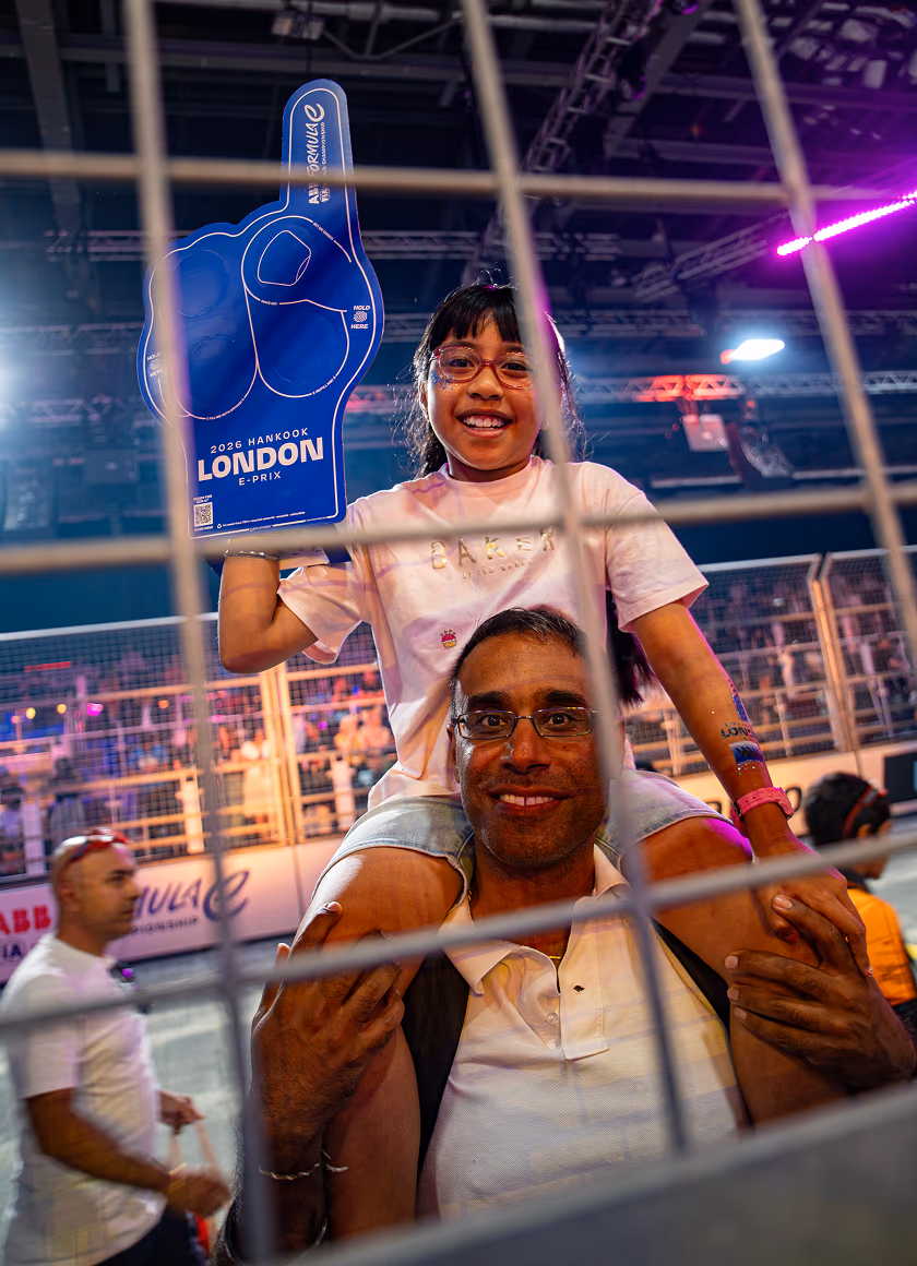 A young girl sitting on a man's shoulders holds up a giant blue foam finger promoting the 2026 Hankook London E-Prix at a Formula E racing event, viewed through a metal fence.
