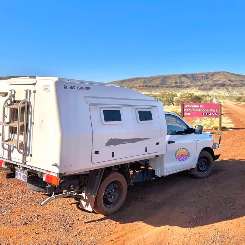 White Rhino Campers vehicle parked on red dirt road near a sign welcoming visitors to Karijini National Park, with a clear blue sky and rocky hills in the background.