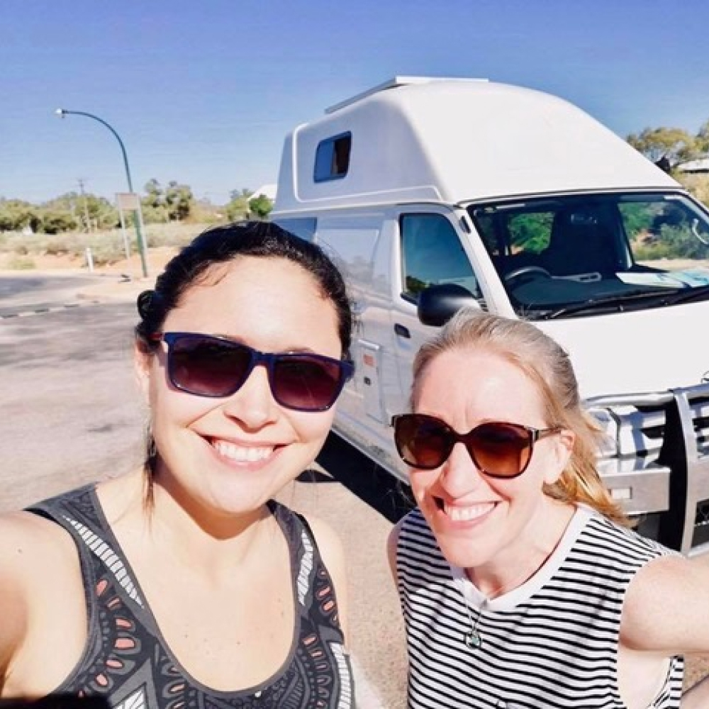 Two women wearing sunglasses smiling and taking a selfie in front of a white camper van on a sunny day.