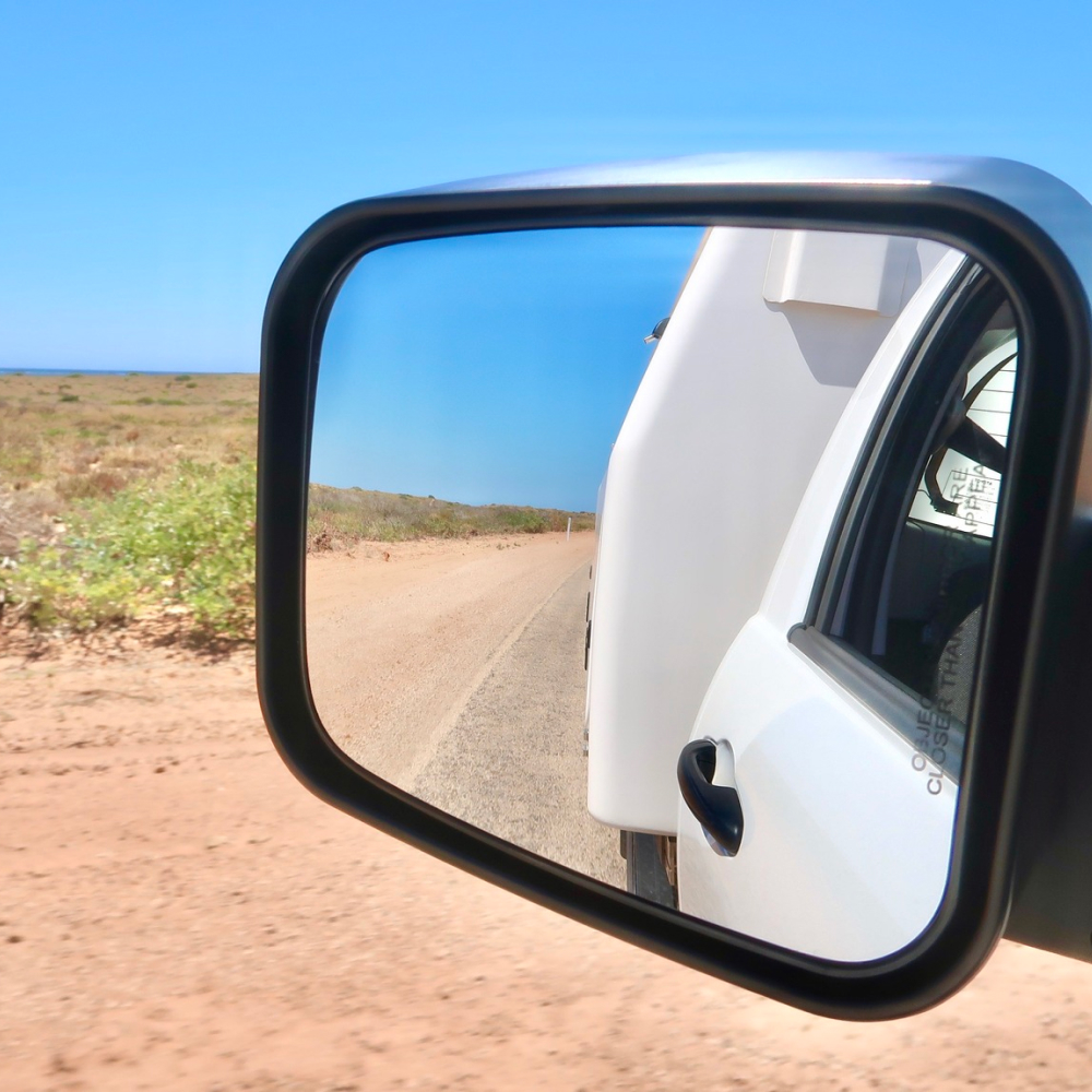 View of a dirt road and dry shrubland reflected in the side mirror of a white vehicle.