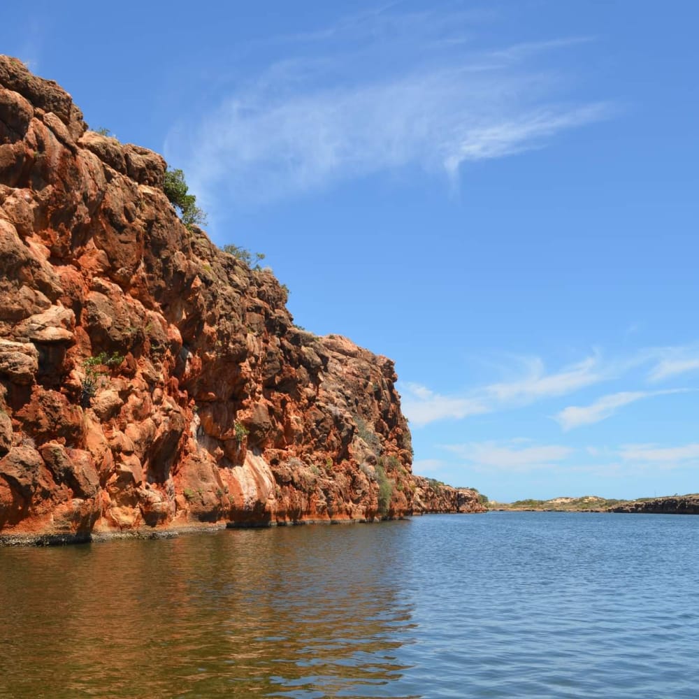 Red rocky cliffs beside calm blue water under a clear sky with scattered clouds.