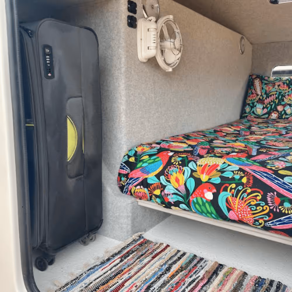 Interior of camper van showing a bed with colourful tropical bird and flower patterned bedding next to upright black suitcase with wheels and a small white wall-mounted fan.