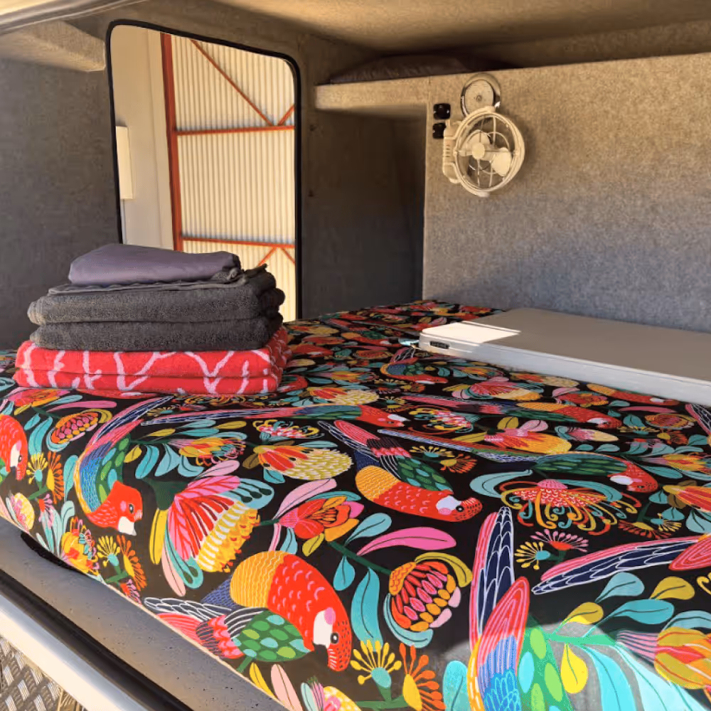 Bed with colorful bird-patterned bedding, folded towels, and a closed laptop inside a compact camper or camper shell.