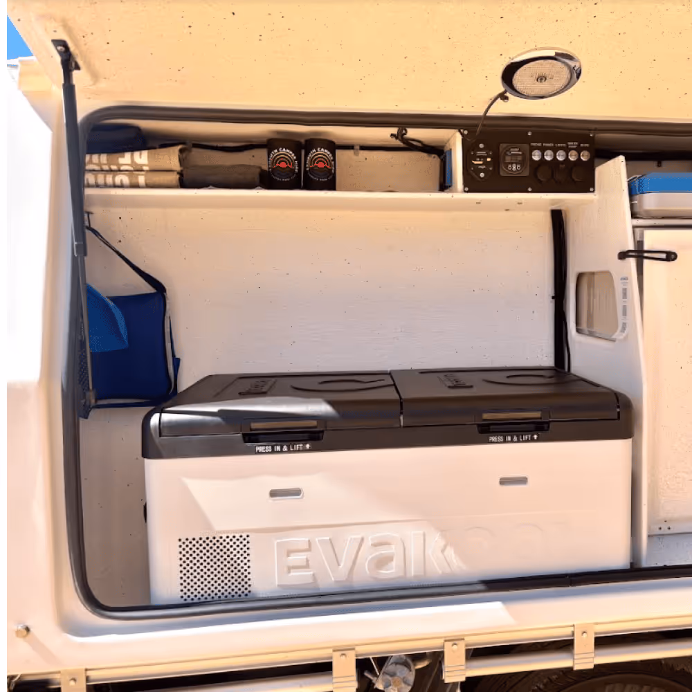 Open vehicle storage compartment with a large white Evakool portable fridge, folded towels, two cans, and a control panel with a speaker above.