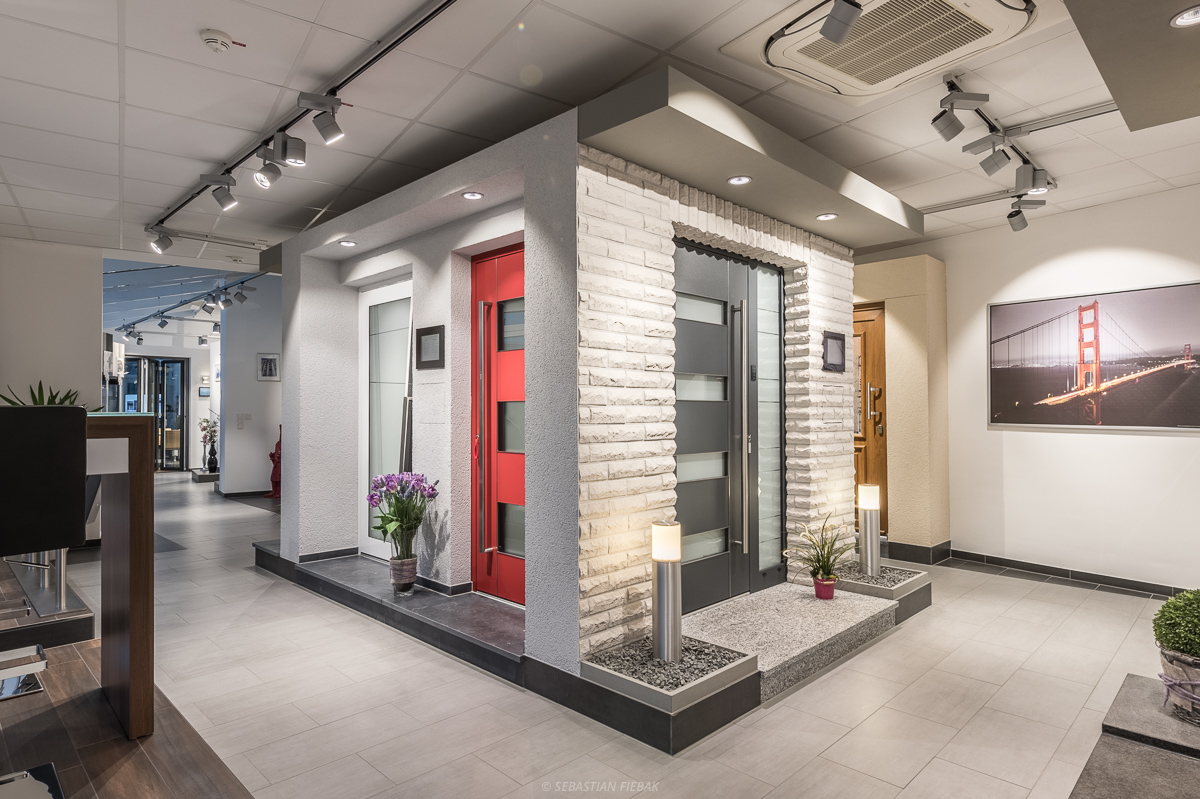 Ausstellungsraum mit modernen Türdisplays, in denen verschiedene Eingangstürstile und -materialien präsentiert werden. Der Raum ist gut beleuchtet und verfügt über Dekorelemente wie Pflanzen und gerahmte Fotos.