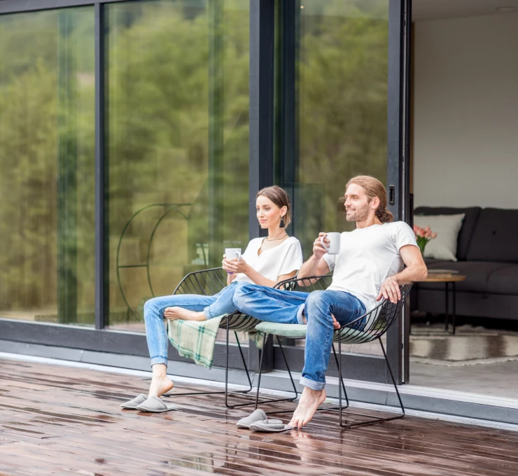 Zwei Personen sitzen entspannt auf Stühlen auf einer Holzterrasse vor großen Glasfenstern und trinken aus Tassen.
