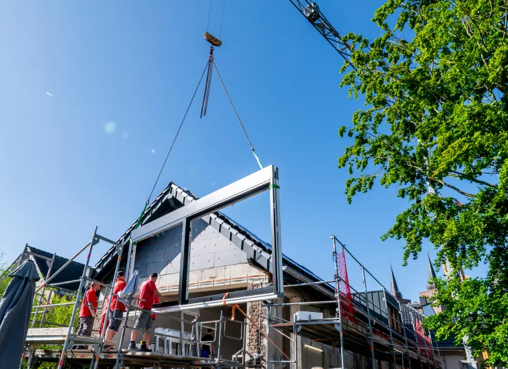Drei Bauarbeiter montieren mit einem Kran ein großes Fenster in ein Haus unter blauem Himmel.