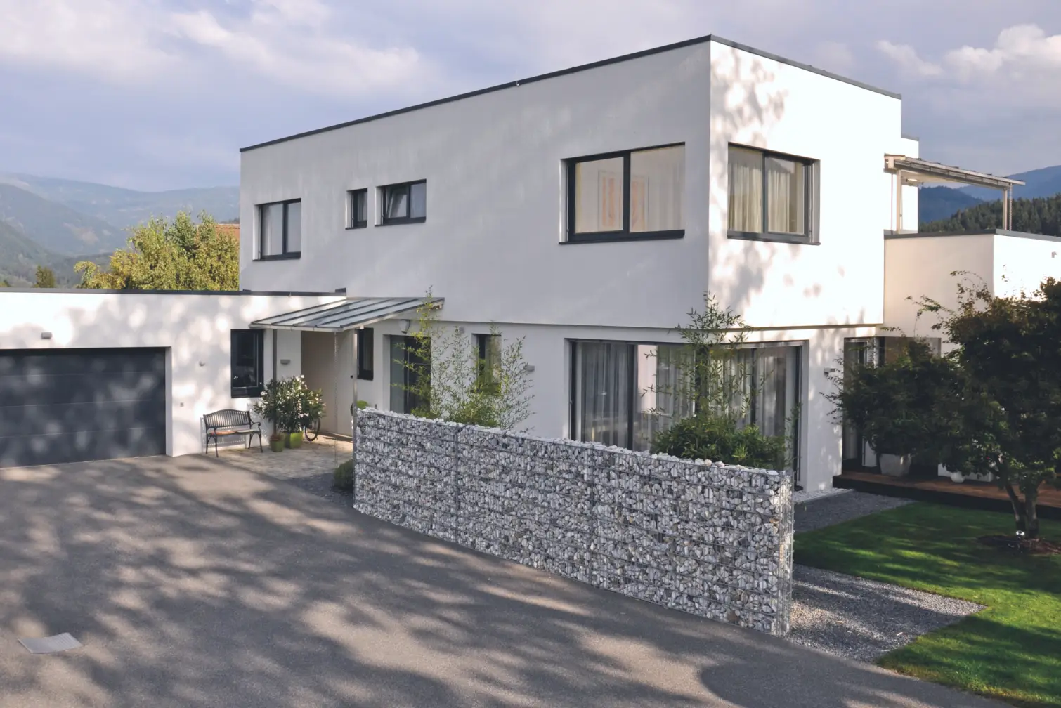 Modernes weißes zweistöckiges Wohnhaus mit großen Fenstern, einer Garage, einer Steinmauer und einem kleinen Garten.