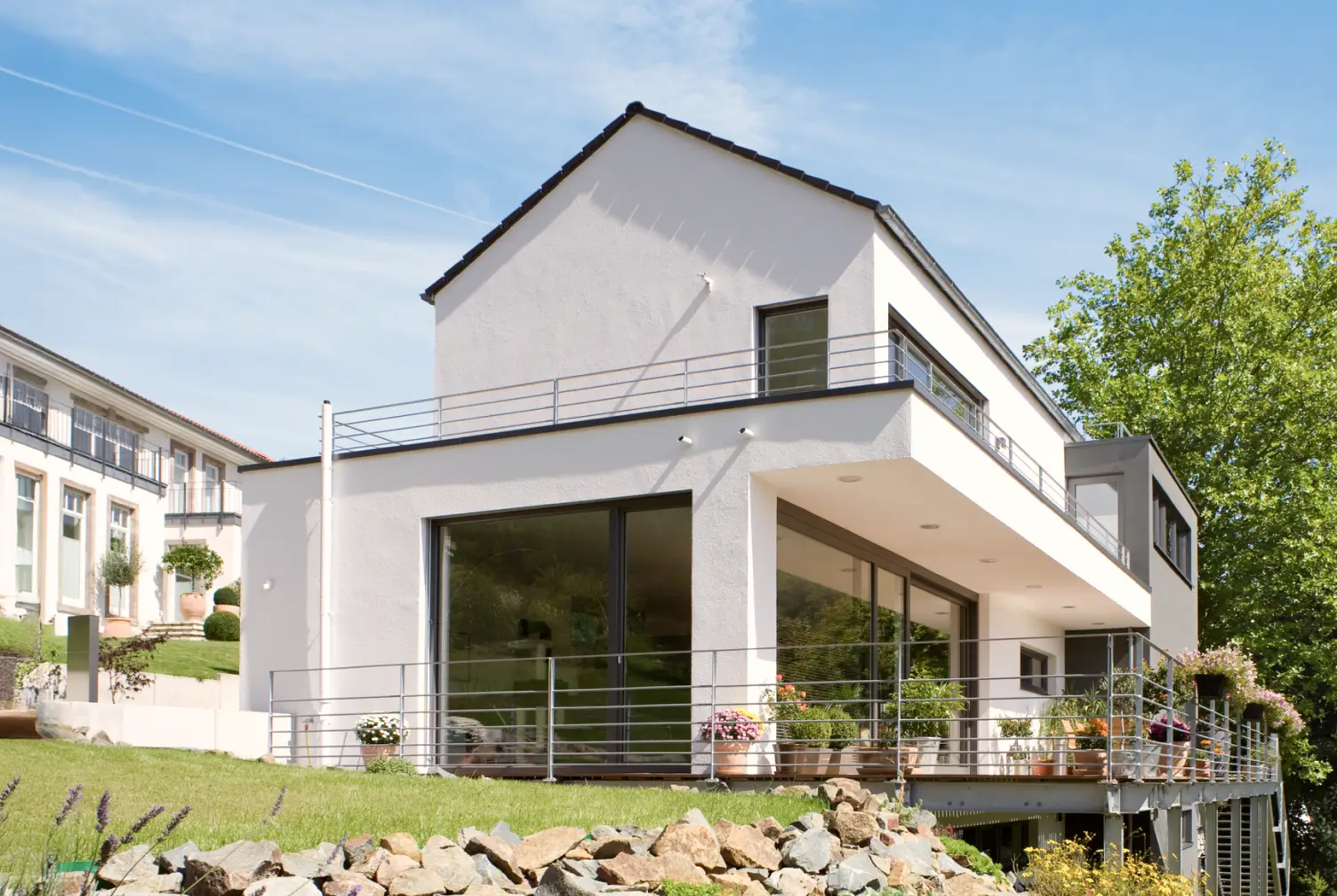 Modernes weißes Einfamilienhaus mit großem Balkon, Glasfronten und umlaufendem Geländer vor blauem Himmel.