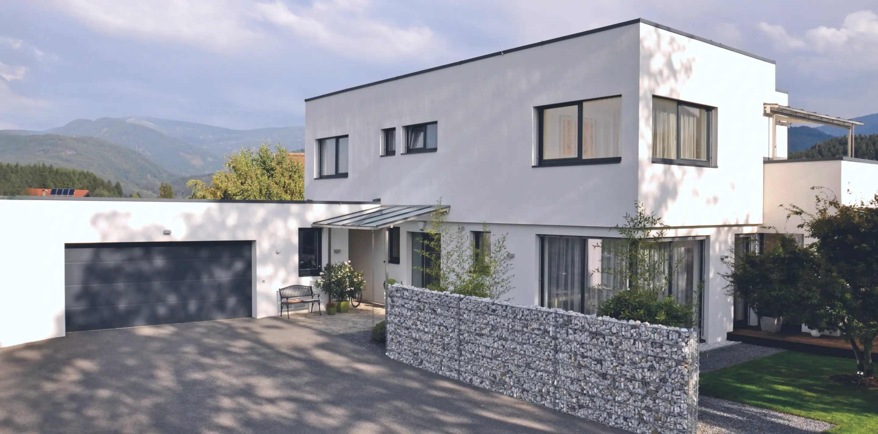 Modernes zweistöckiges weißes Haus mit Garage, Glasvordach und Steingabionenzaun vor bergiger Landschaft.