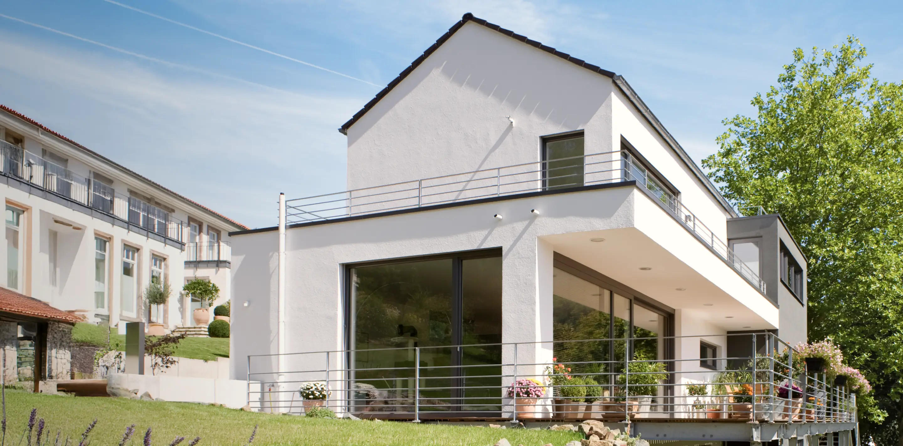 Modernes weißes Haus mit großen Fenstern, Terrasse mit Pflanzen und grünem Garten unter blauem Himmel.