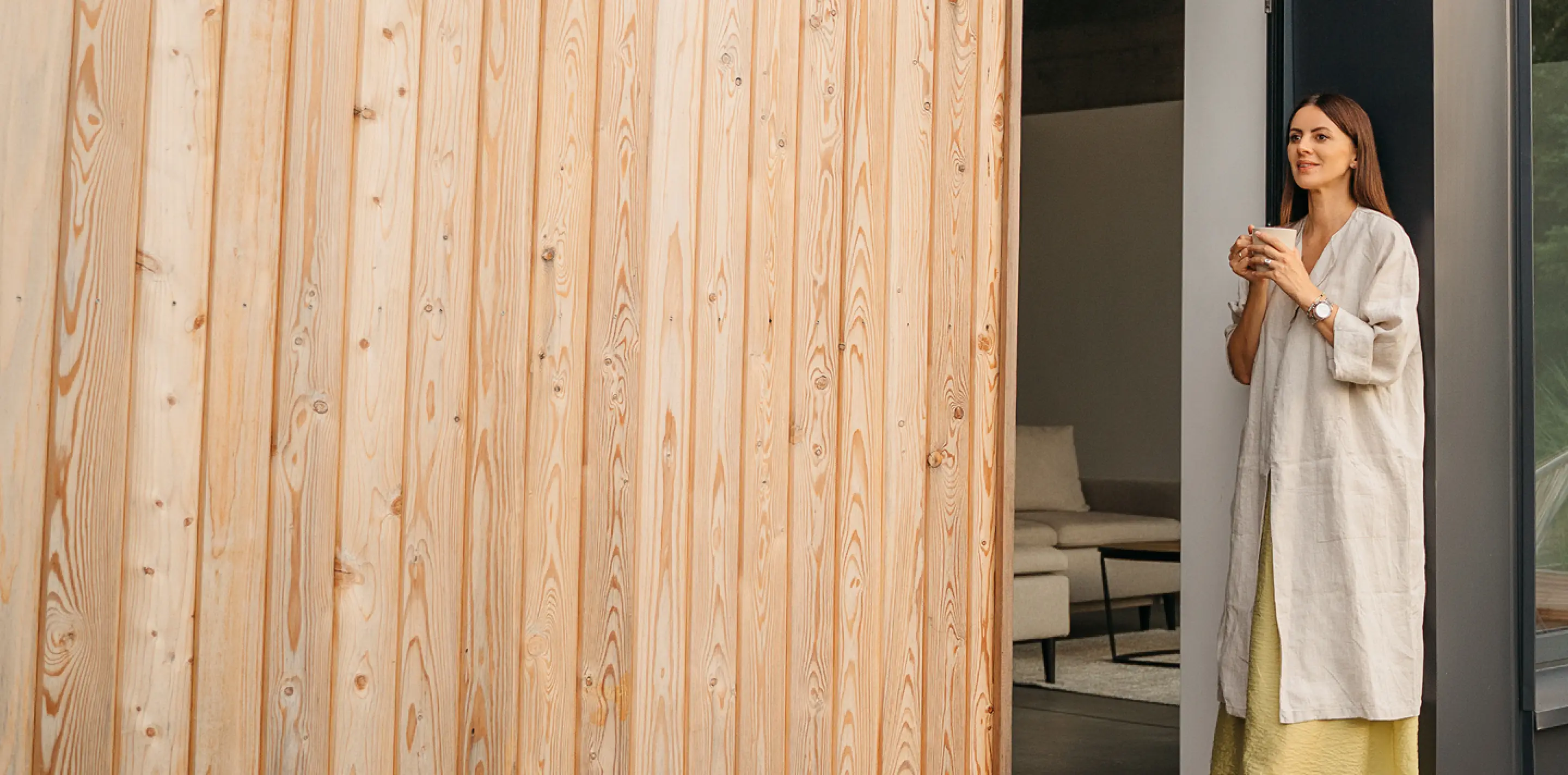 Frau steht lächelnd in einer Türöffnung eines modernen Hauses und hält eine Tasse.