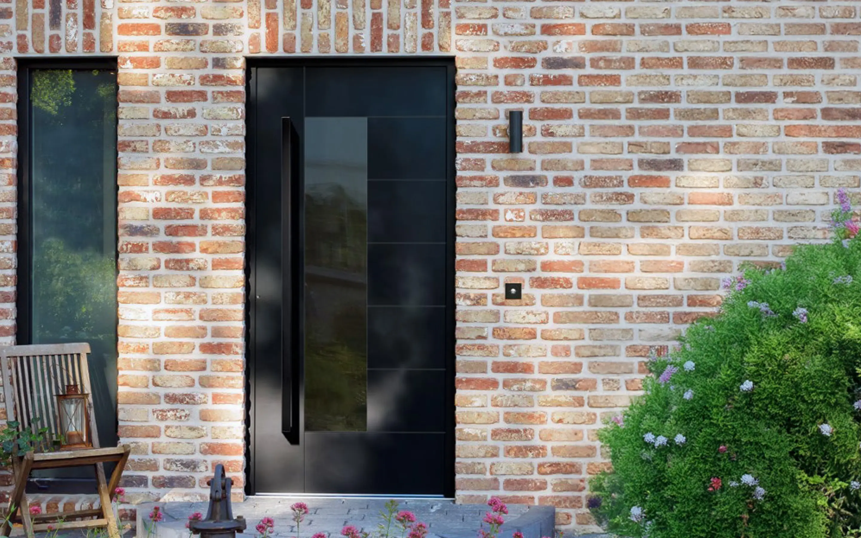 Eingangstür aus schwarzen Paneelen mit großem Griff an einer Ziegelsteinwand, daneben Holzstuhl mit Laterne und blühende grüne Büsche.