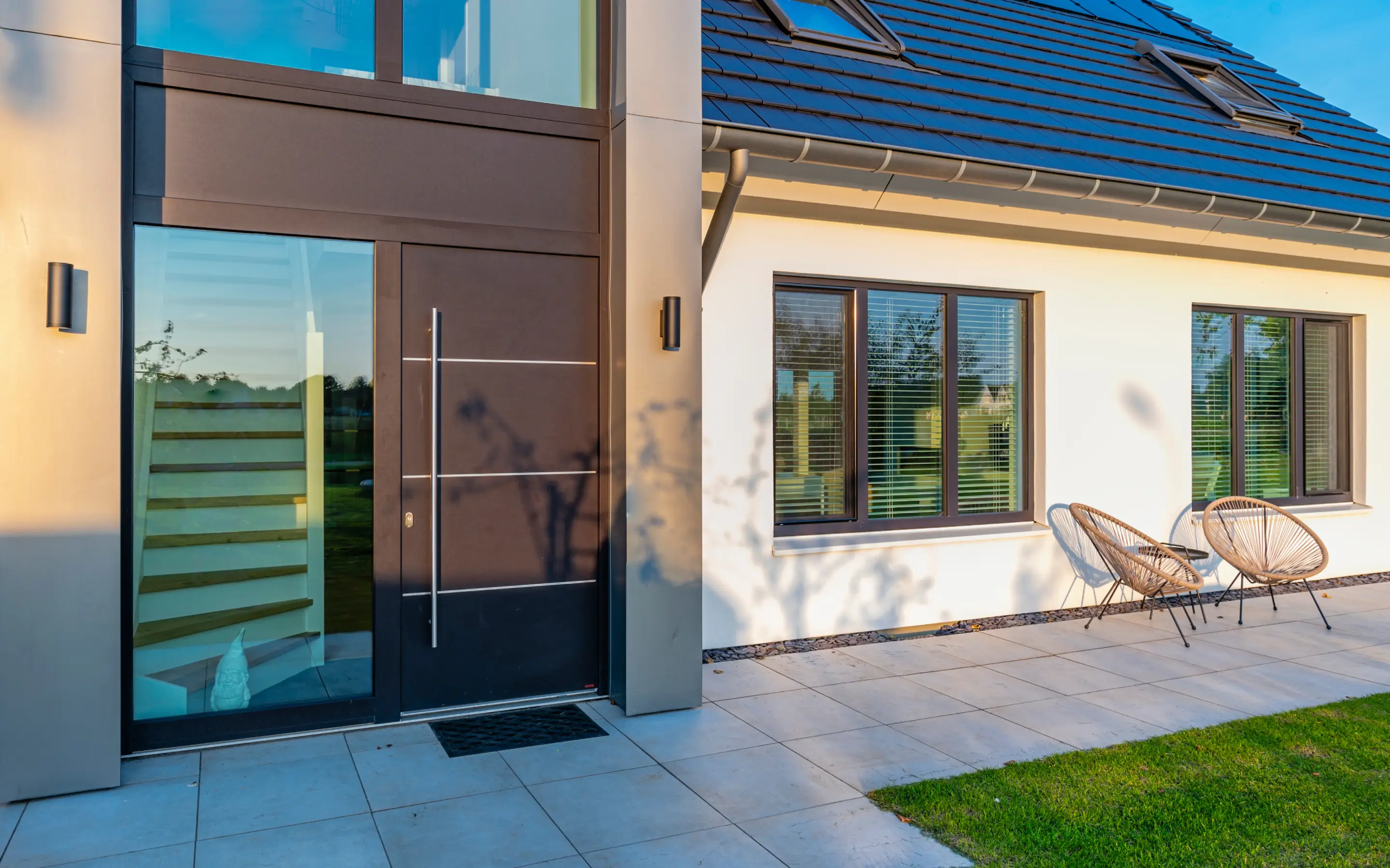 Moderner Hauseingang mit dunkler Tür, großen Fenstern, zwei Stühlen und einem kleinen Tisch auf der Terrasse.