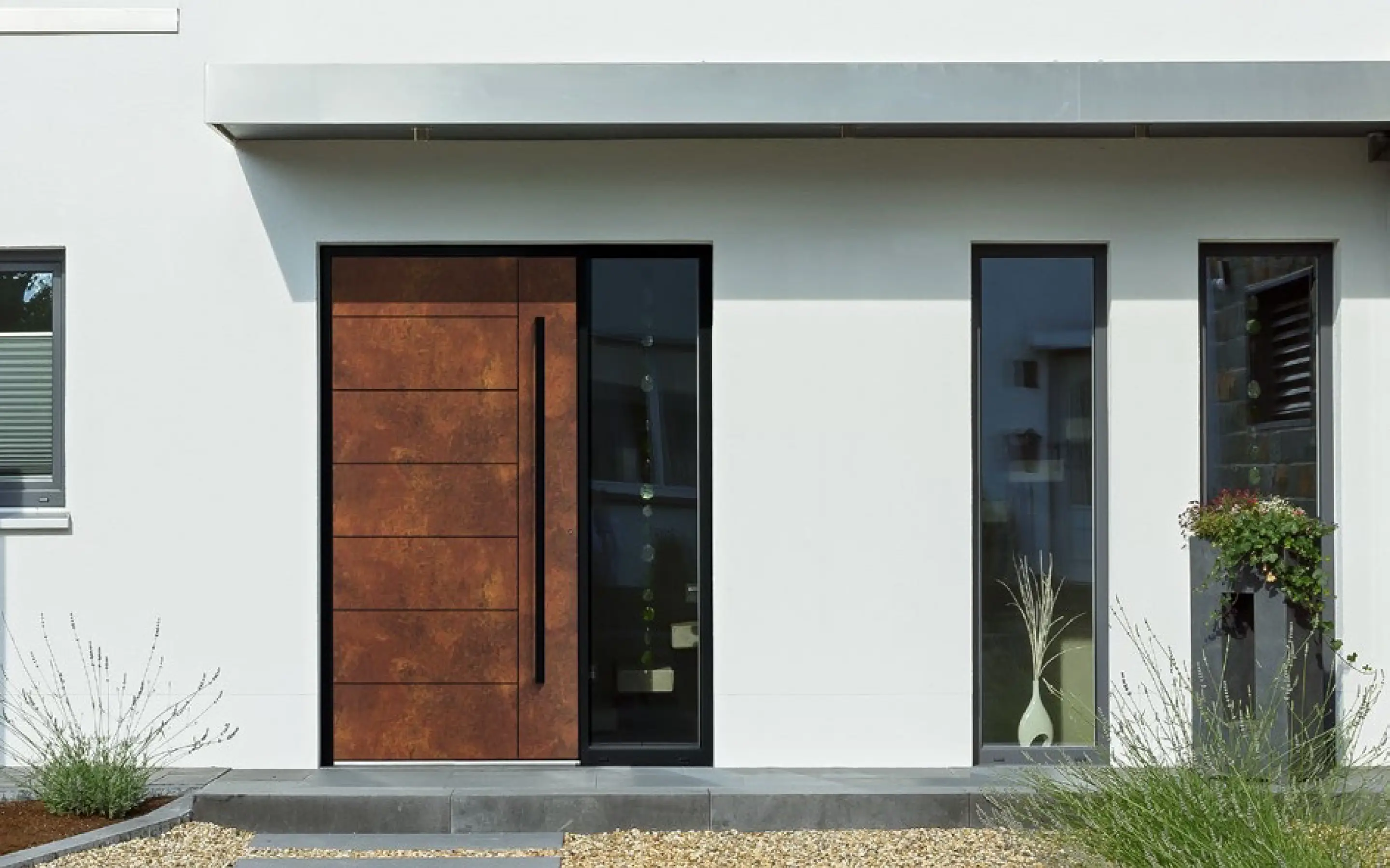 Moderner Hauseingang mit einer großen braunen Holztür, zwei schlanken Fenstern und weißer Fassade.