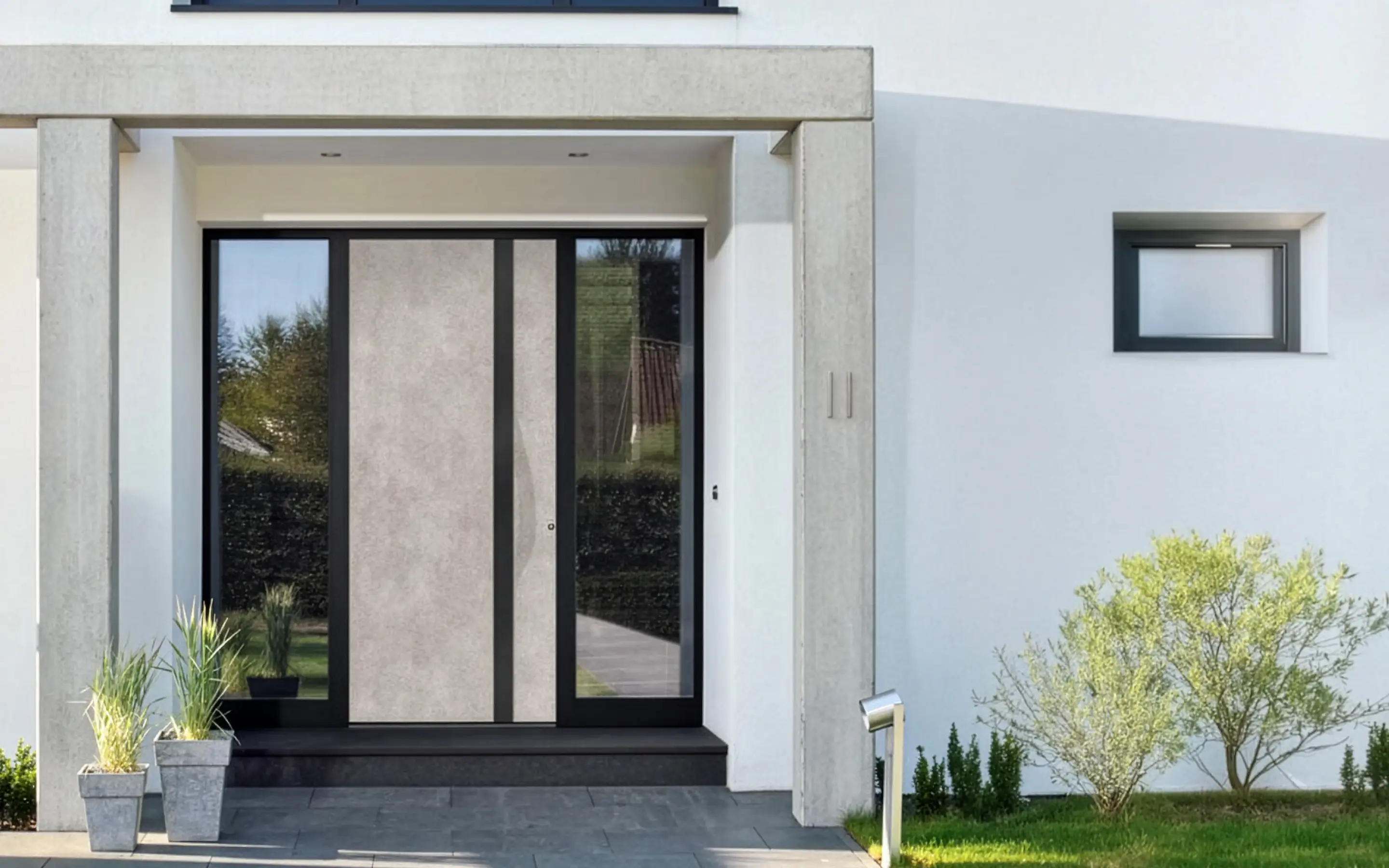 Moderner Hauseingang mit grauer Tür, Fußweg aus dunklen Platten und kleinen Pflanzen in Töpfen links, sowie Busch auf grüner Wiese rechts.
