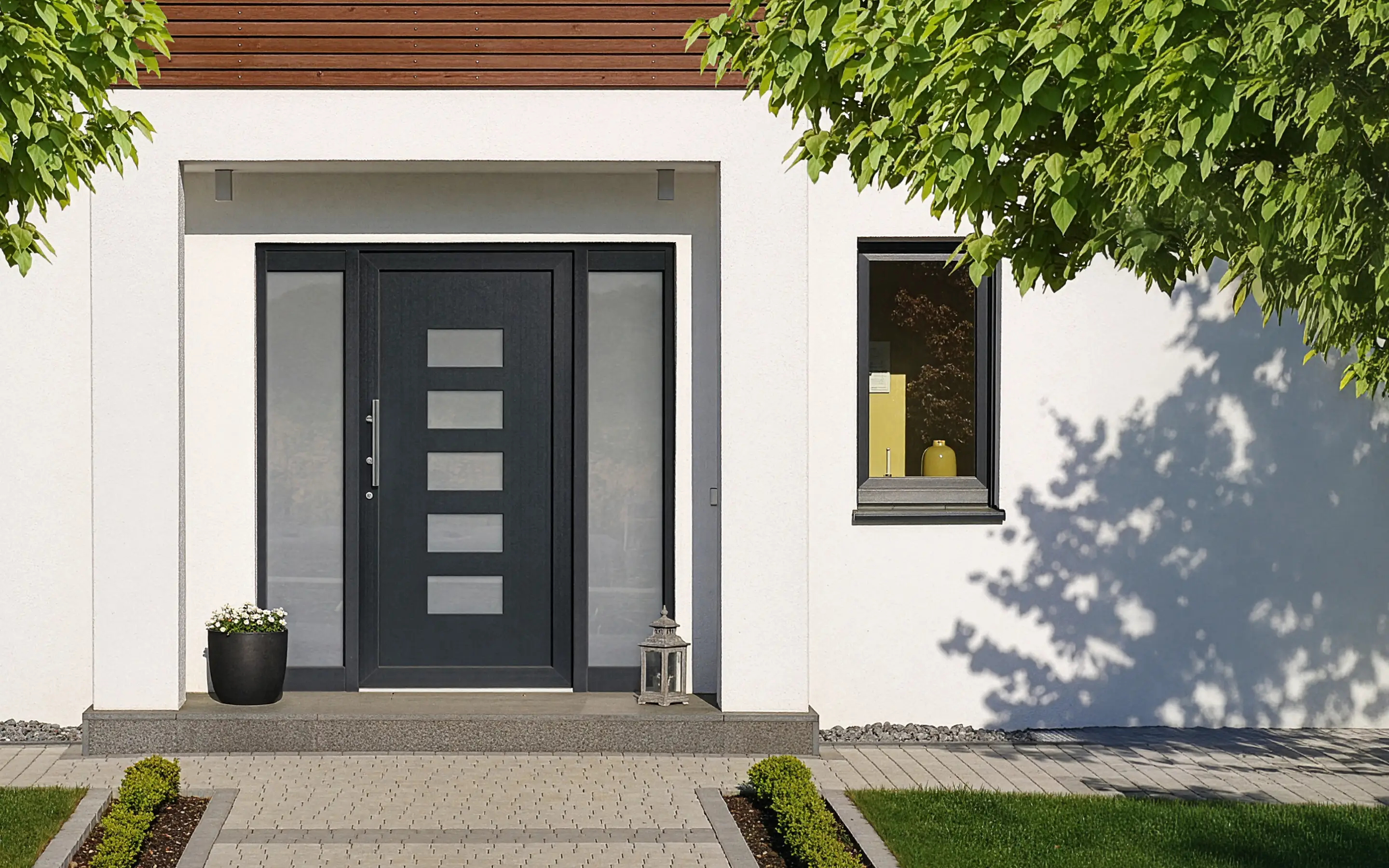 Moderner Hauseingang mit schwarzer Tür, Blumenkübel und Laterne, umgeben von grünem Rasen und Bäumen.