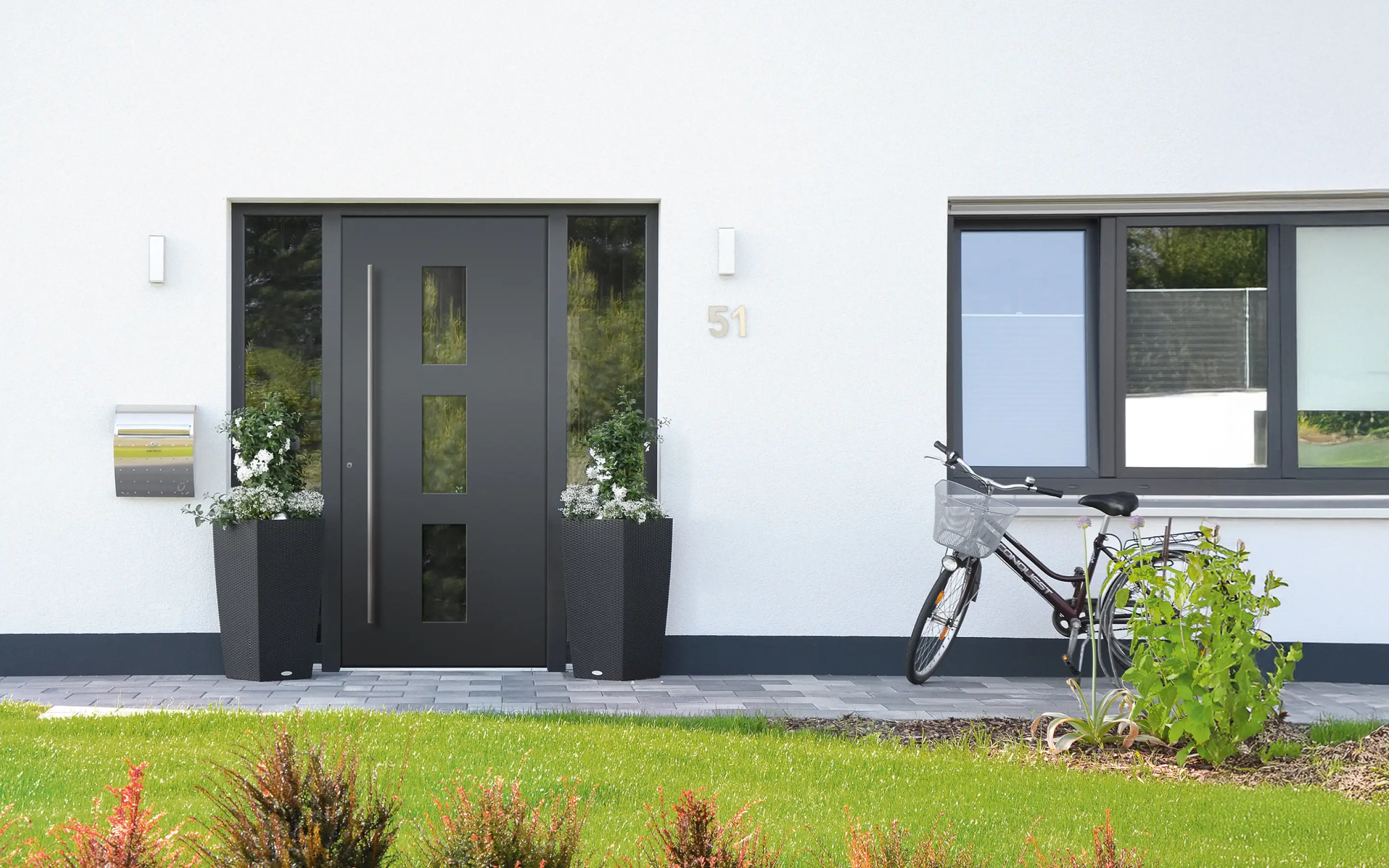 Moderner Hauseingang mit dunkler Tür, zwei Pflanzen in schwarzen Töpfen, Briefkasten und Fahrrad neben einem Fenster.
