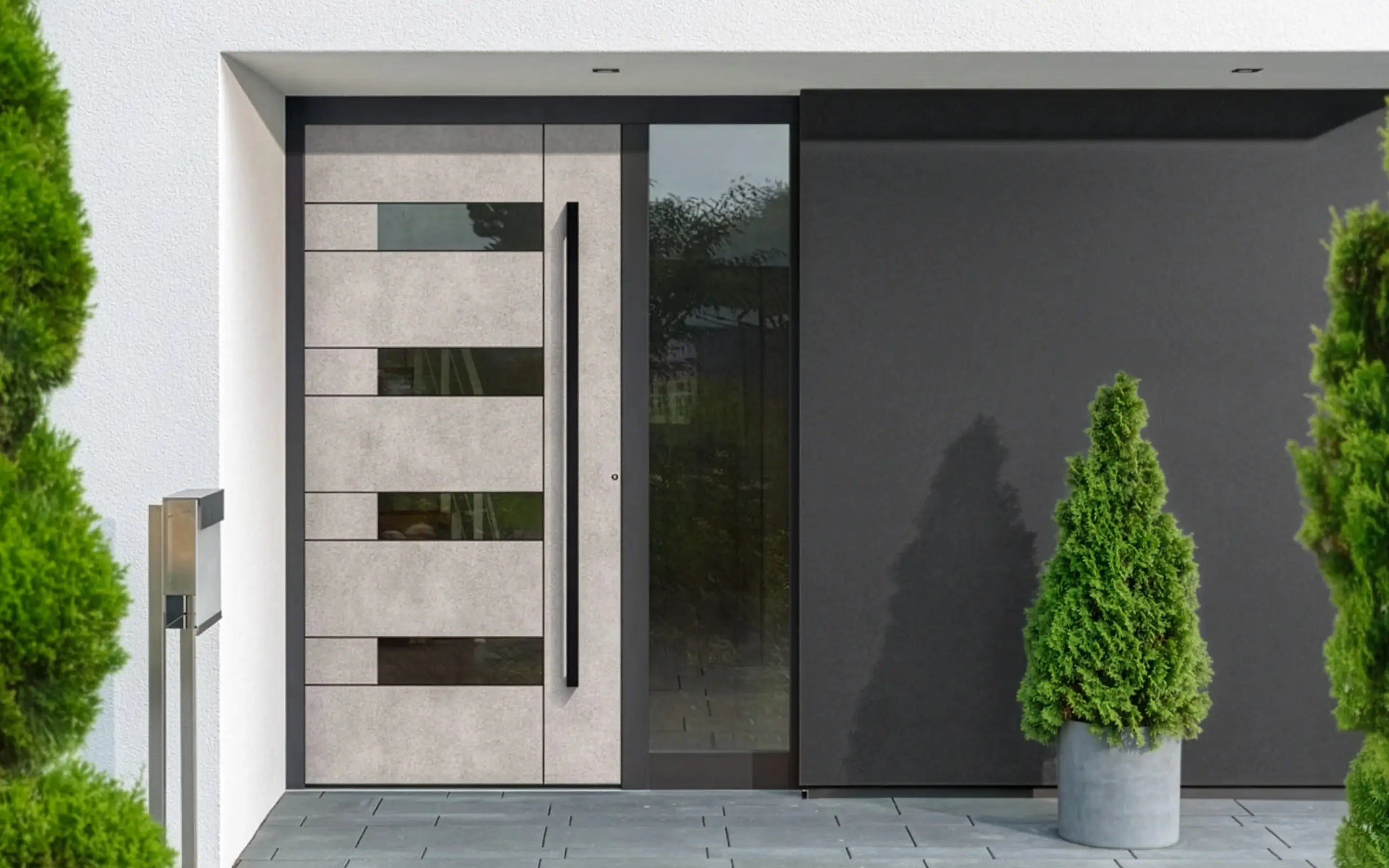 Moderner Hauseingang mit einer hellgrauen Tür mit rechteckigen Glasfenstern und einem schwarzen Griff, umgeben von weißen und dunkelgrauen Wänden, mit grünen Pflanzen in grauen Töpfen außen.