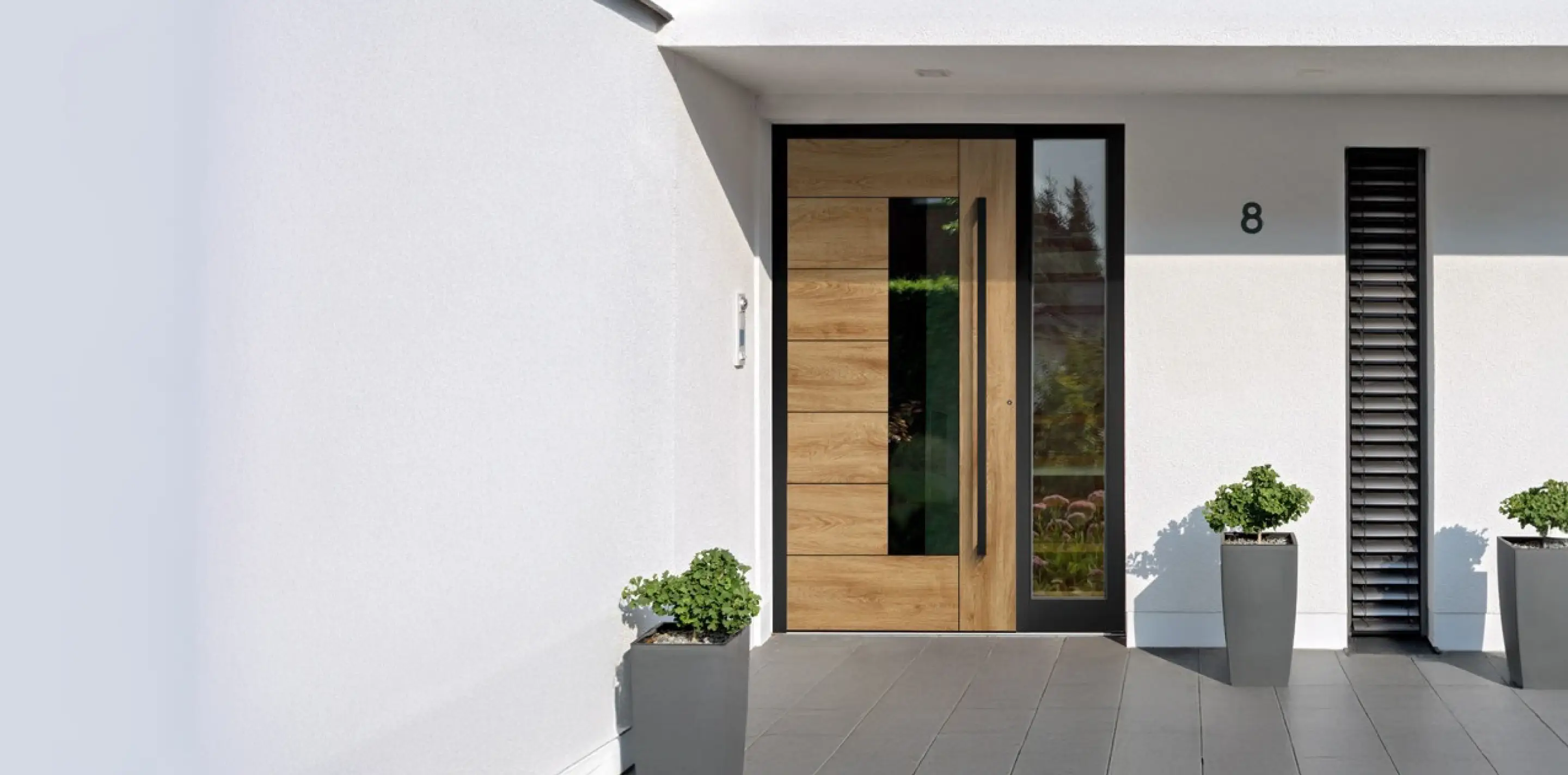 Moderner Hauseingang mit Holztür, seitlichem Glasfenster, drei grauen Pflanzgefäßen mit Grünpflanzen und der Hausnummer 8.