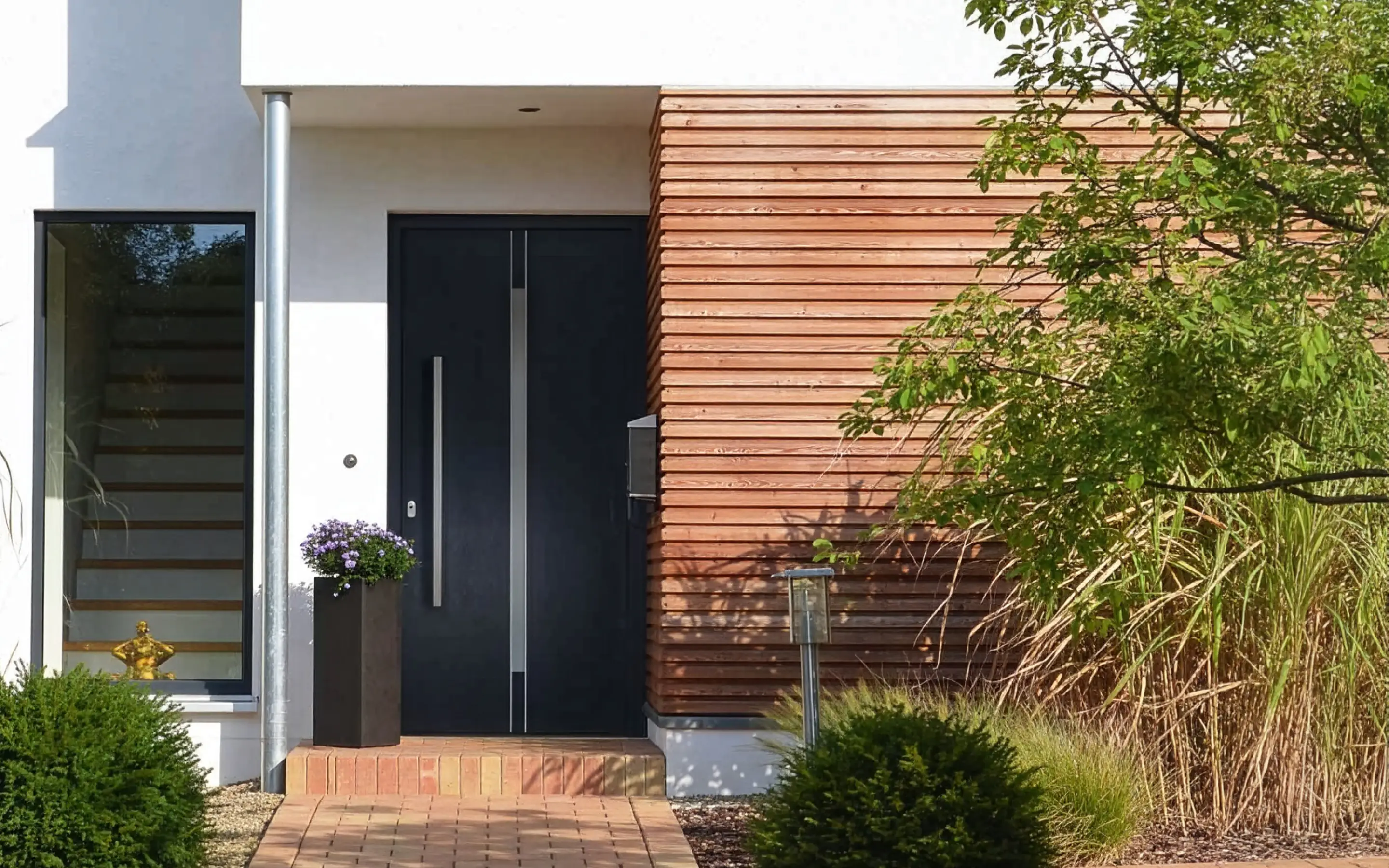 Moderner Hauseingang mit schwarzer Tür, Holzverkleidung neben der Tür und einem Blumenkübel mit lila Blumen.