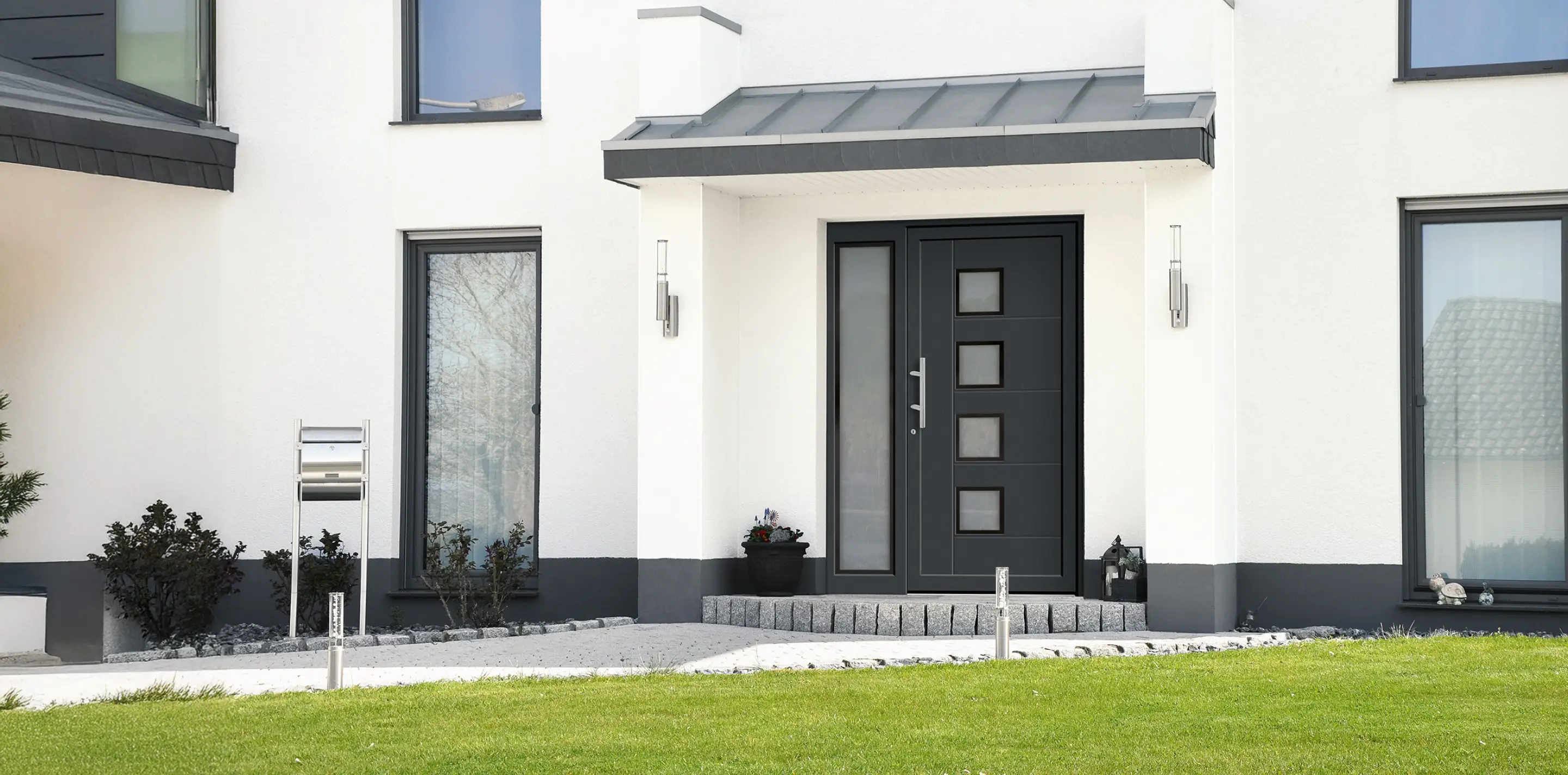 Eingangsbereich eines modernen weißen Hauses mit schwarzer Haustür, vier quadratischen Fenstern und seitlichem Fenster.