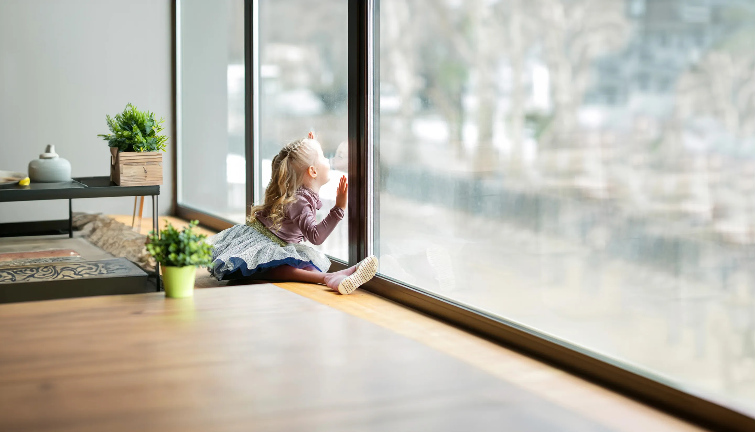 Ein kleines Mädchen sitzt auf dem Boden und schaut neugierig aus einem großen Fenster.