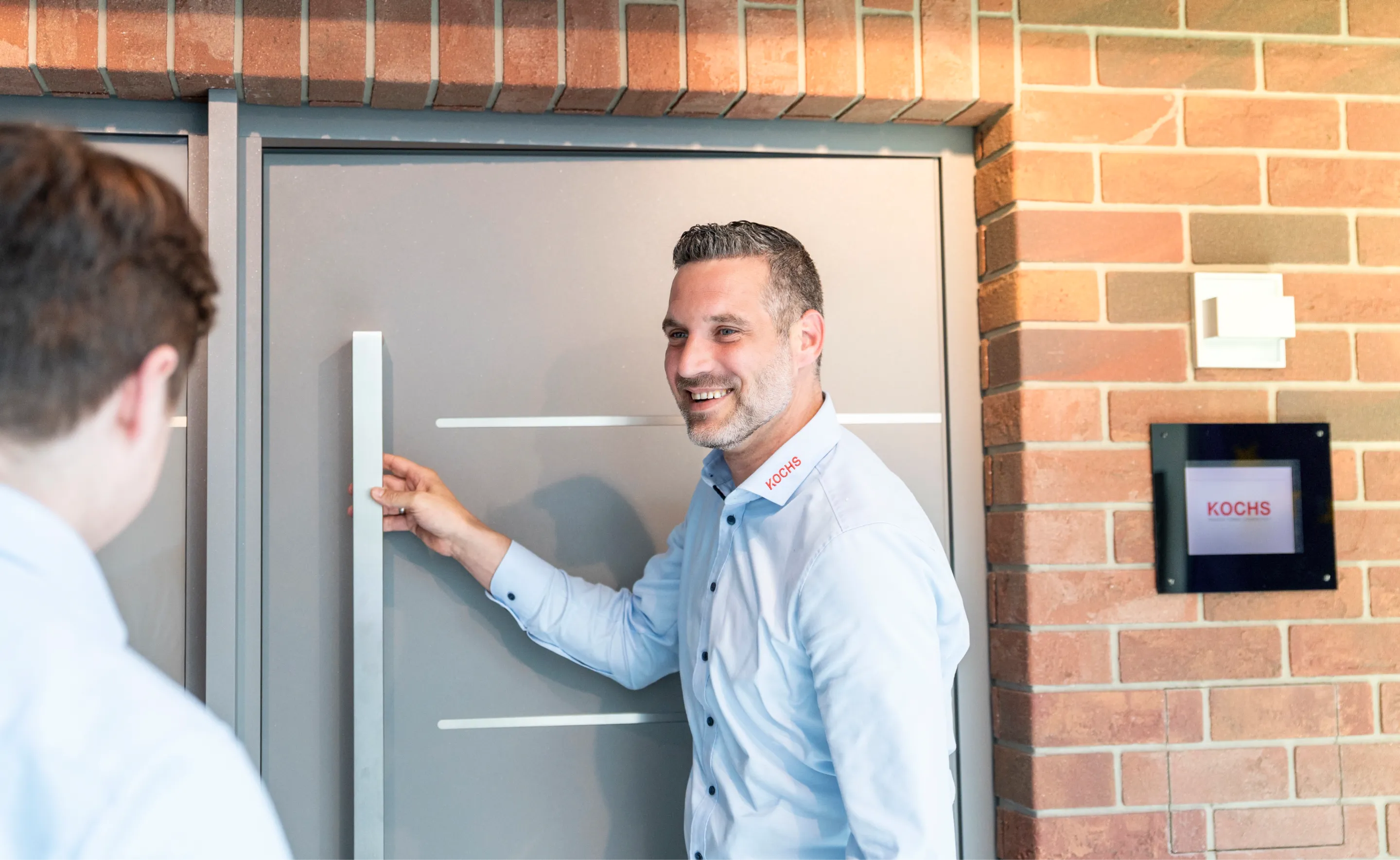 Ein lächelnder Mann in weißem Hemd öffnet eine graue Tür mit einem großen Griff, während eine andere Person im Vordergrund steht.