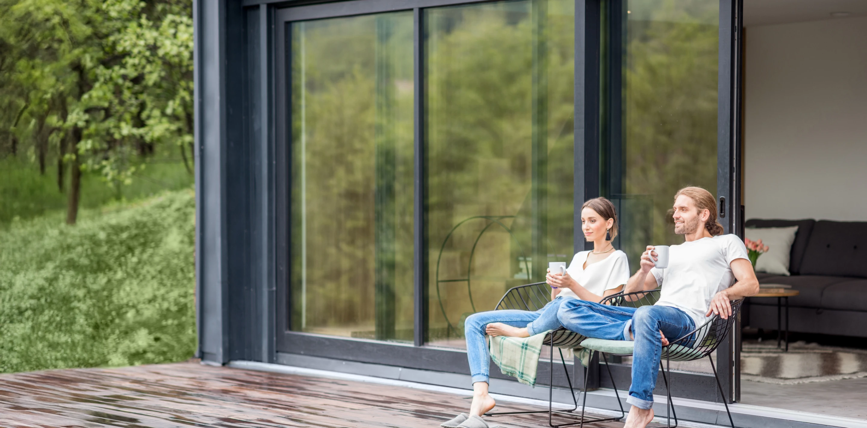 Paar entspannt mit Kaffee auf Stühlen auf der Veranda vor einem modernen Haus mit großem Glasfenster.