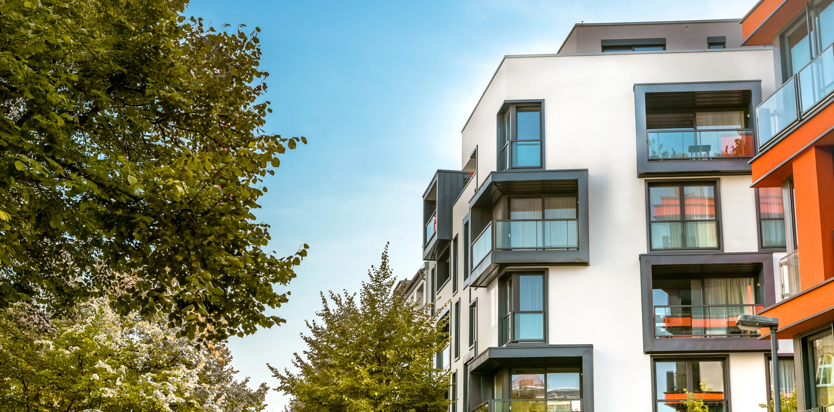 Moderne Wohngebäude mit Balkonen neben grünen Bäumen unter blauem Himmel.